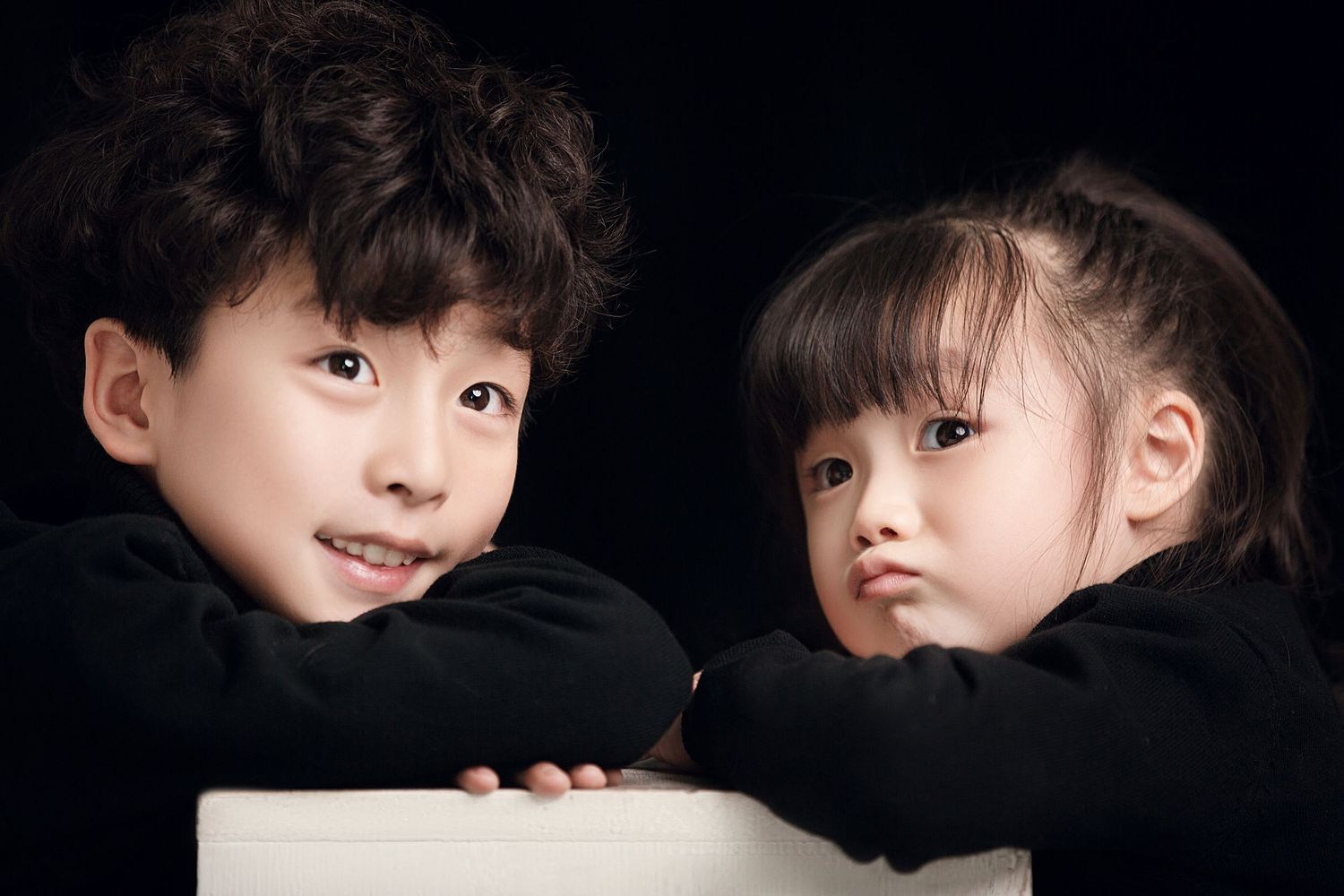 Two Asian children in matching black turtlenecks pose together against a dark studio background.