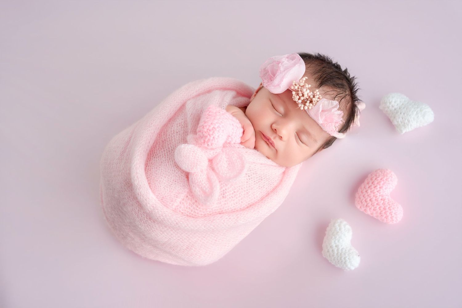 Newborn baby girl sleeping in pink wrap with bow headband, surrounded by knitted hearts on lavender background.