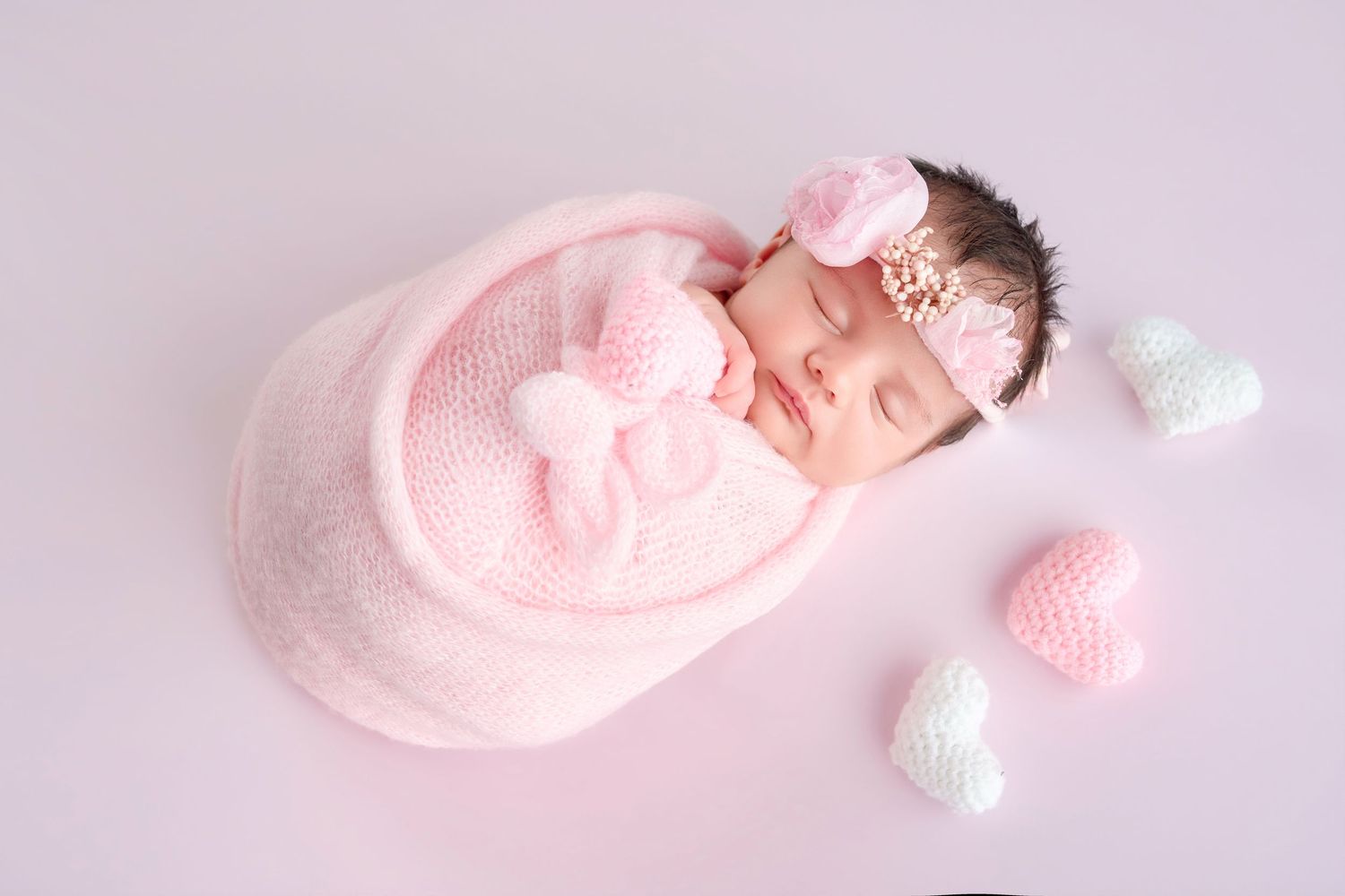 Newborn baby girl sleeping peacefully wrapped in pink knit blanket with heart decorations and floral headband.