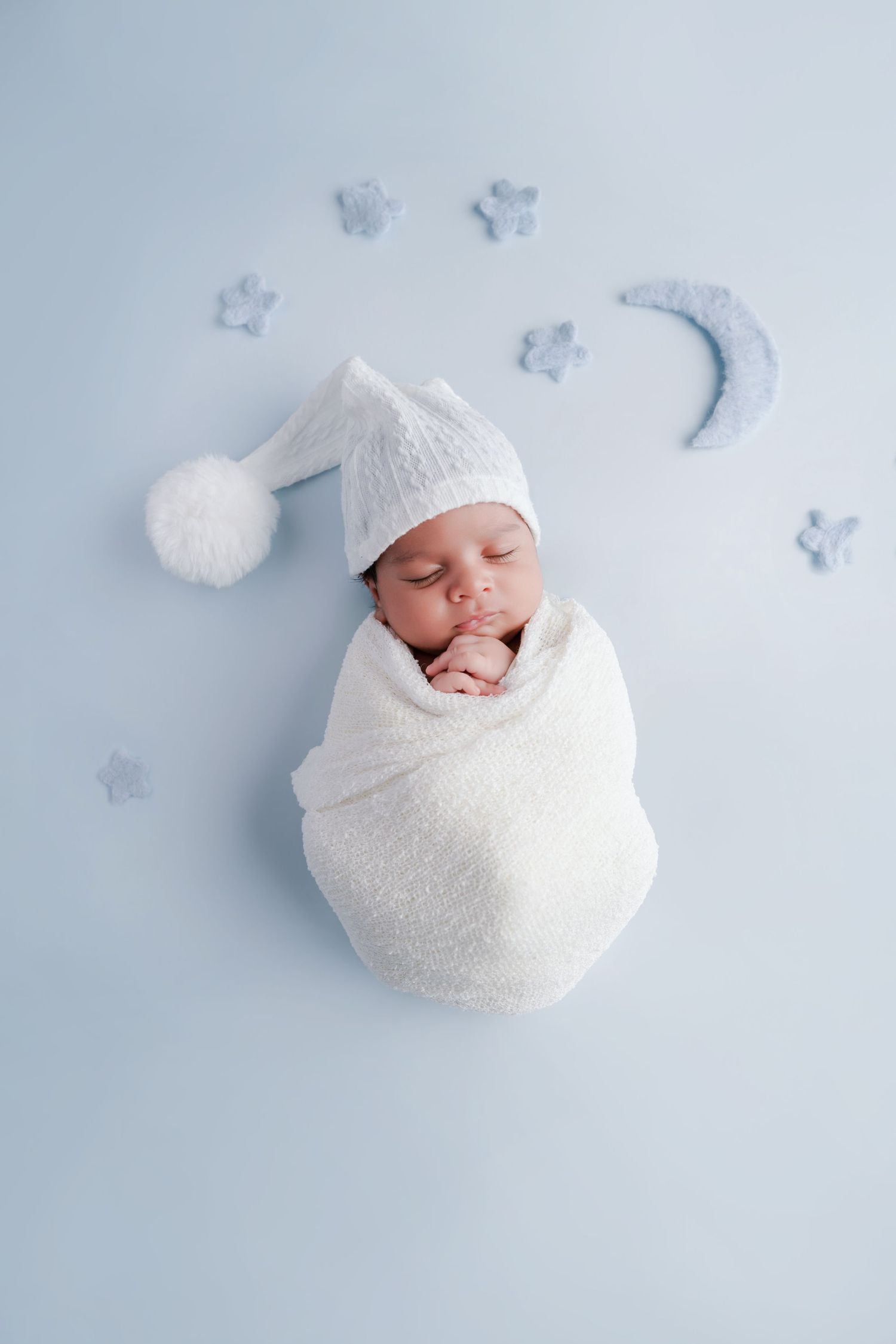 Sleeping newborn baby swaddled in white wrap wearing a knit nightcap, surrounded by stars and moon on blue background.