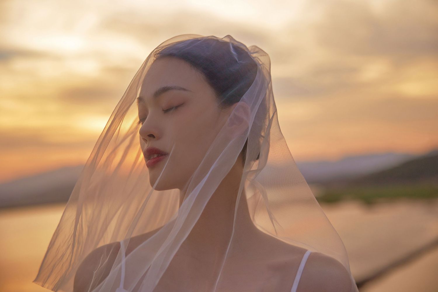 Close-up of bride in white veil with eyes closed at a glowing sunset.