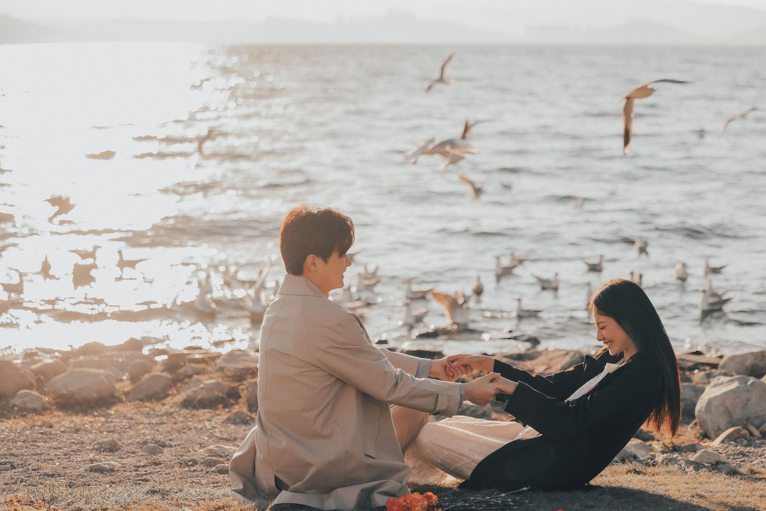 A couple sits on a rocky shore holding hands with seagulls flying over the water behind them.