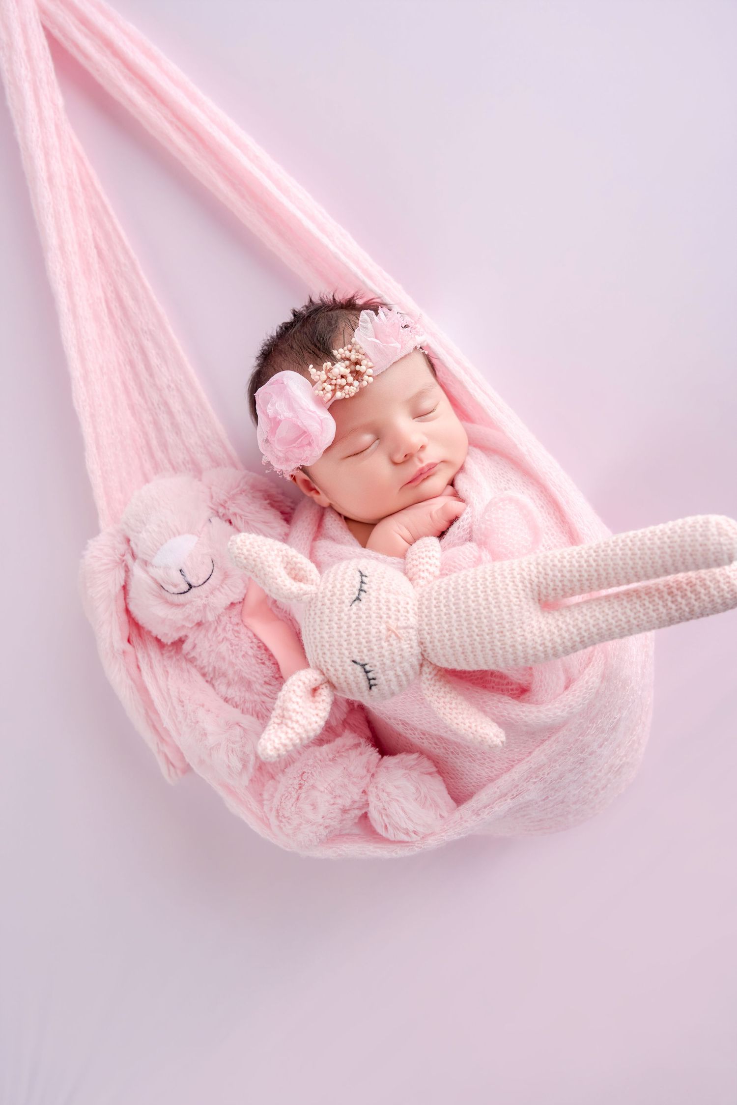 Sleeping newborn baby girl in pink hammock with crochet bunny toys, wearing floral headband.
