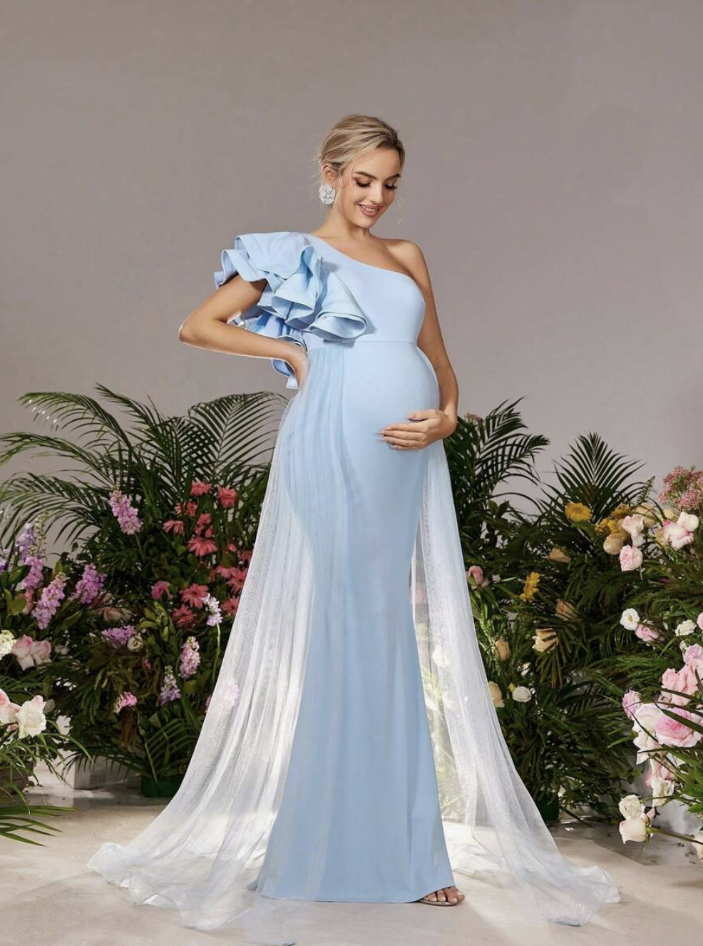 Pregnant woman in a light blue one-shoulder ruffle maternity gown with sheer cape train, posed among lush greenery.