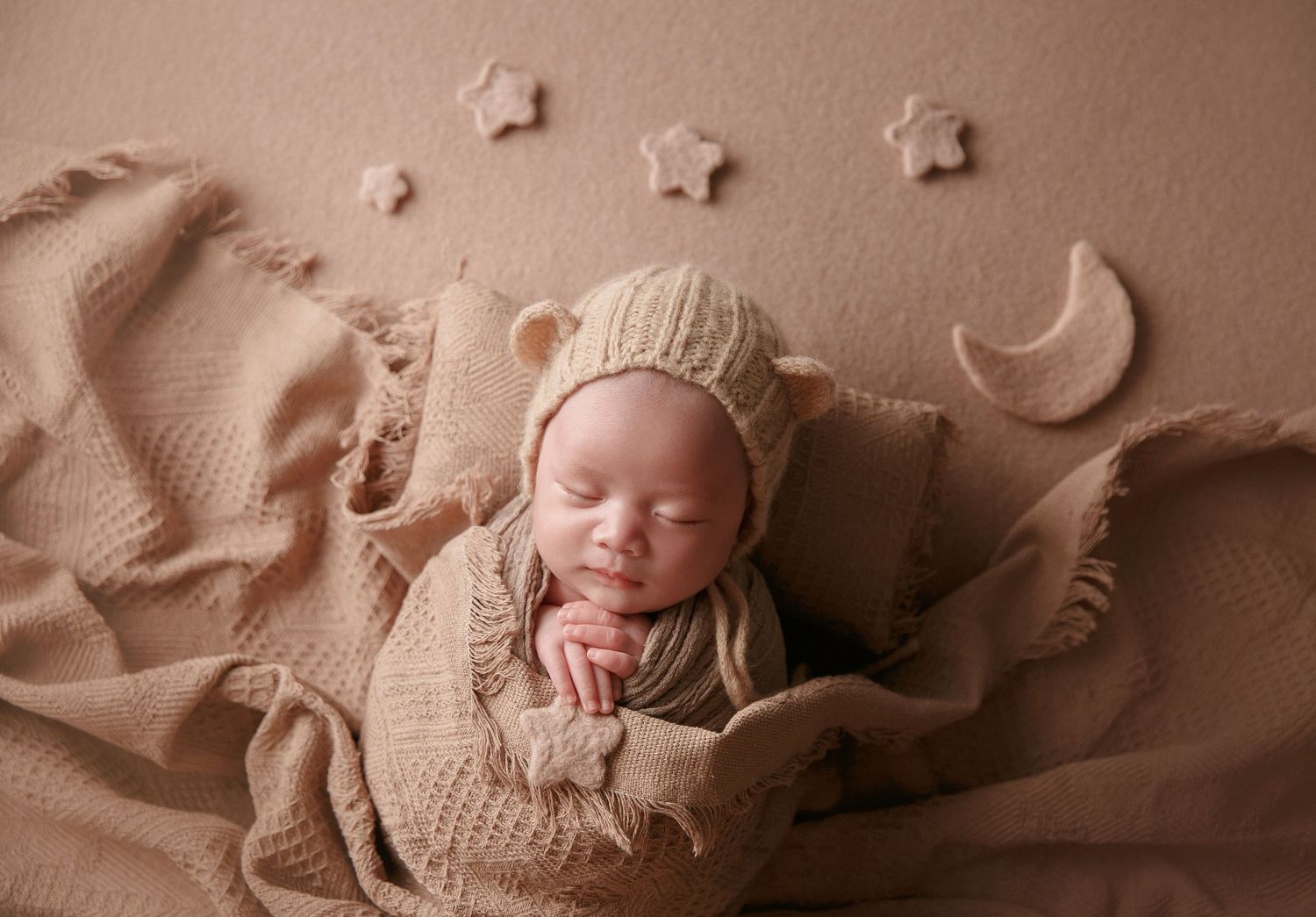 Newborn baby in bear ear bonnet sleeps peacefully on beige blanket with moon and star props.