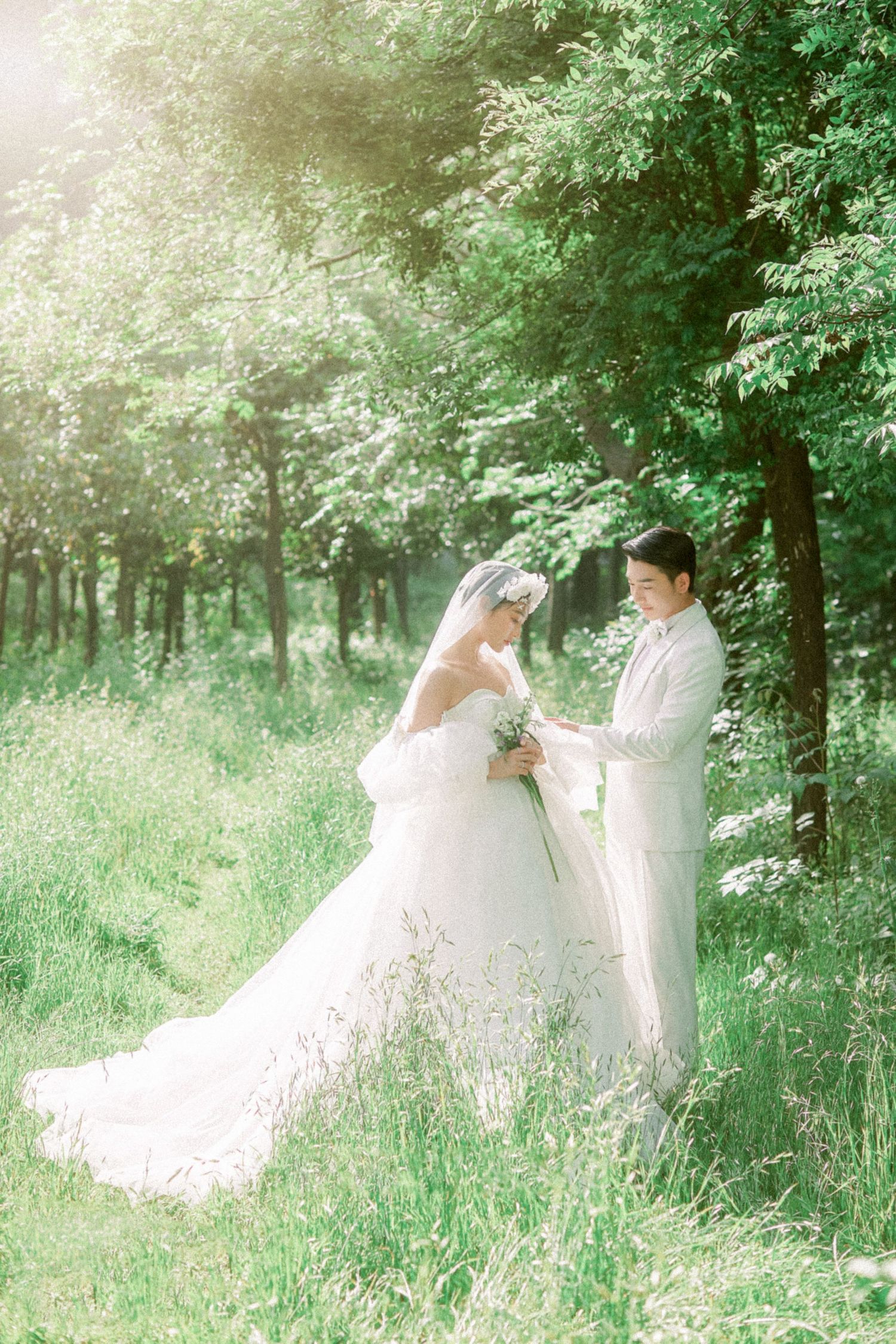 Bride in white ball gown and groom in white suit share a romantic moment in a sunlit green forest.