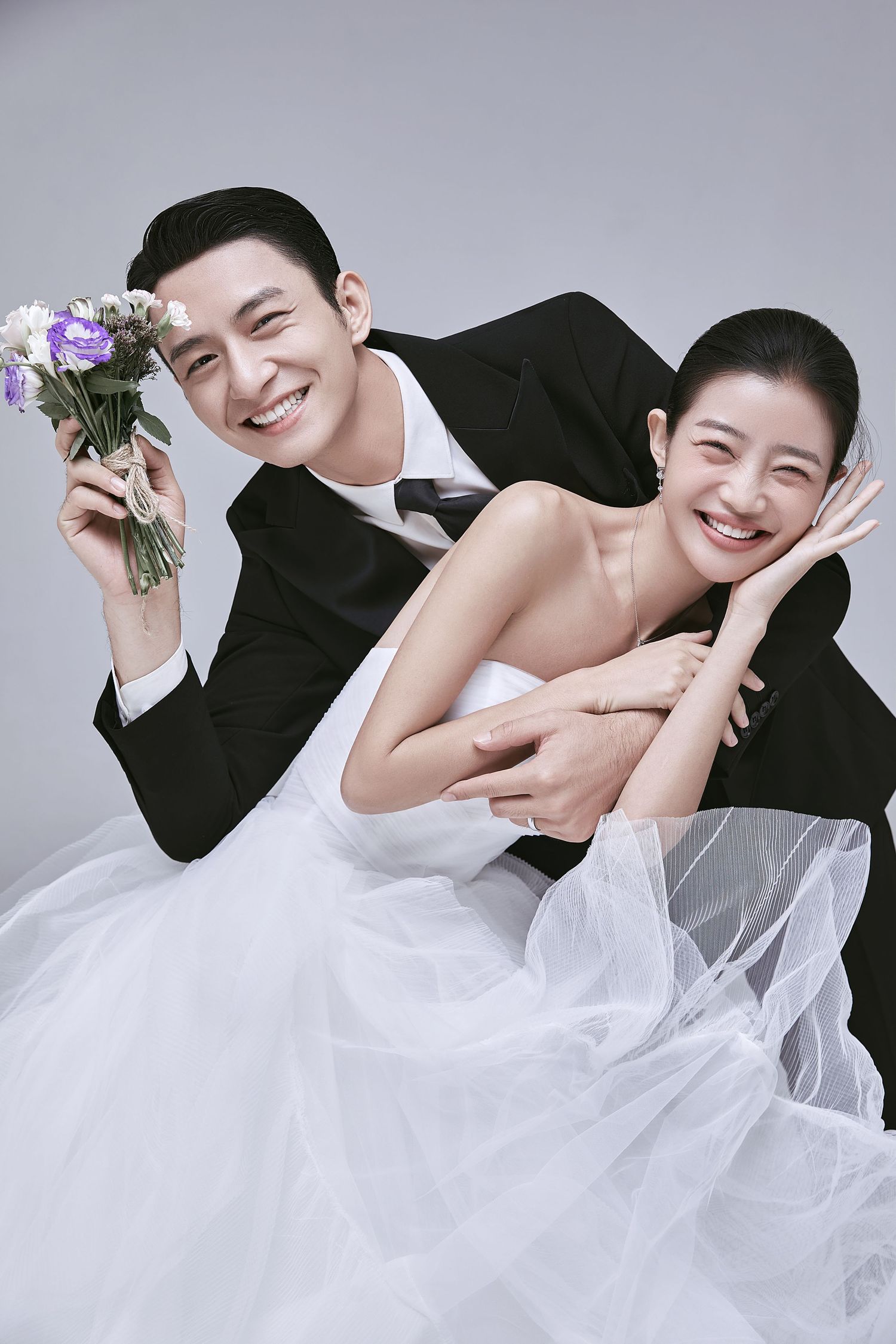 Smiling Asian wedding couple posing playfully, groom holding purple bouquet in studio portrait.