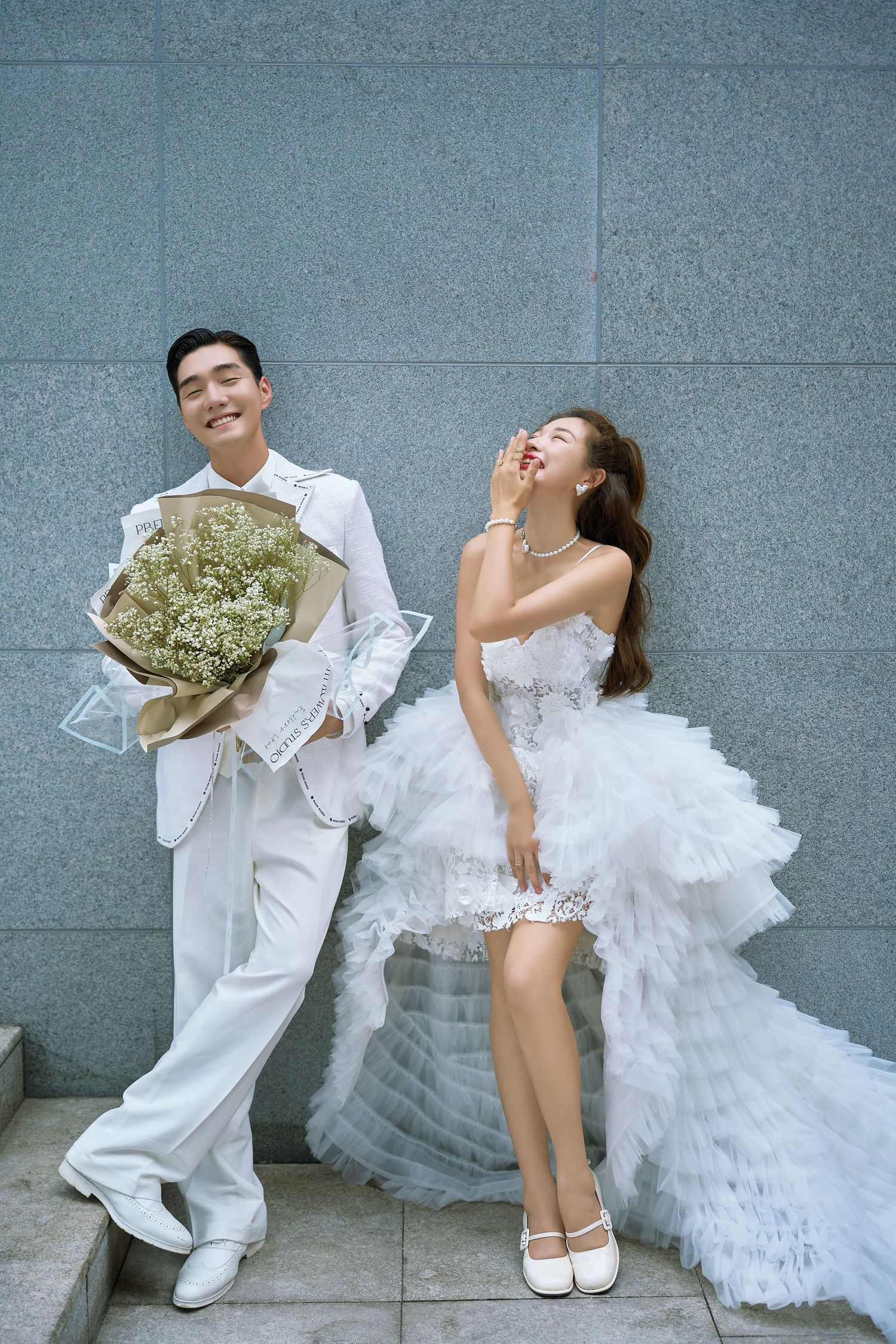 Stylish wedding couple in white outfits posing against gray wall, groom holding large bouquet.
