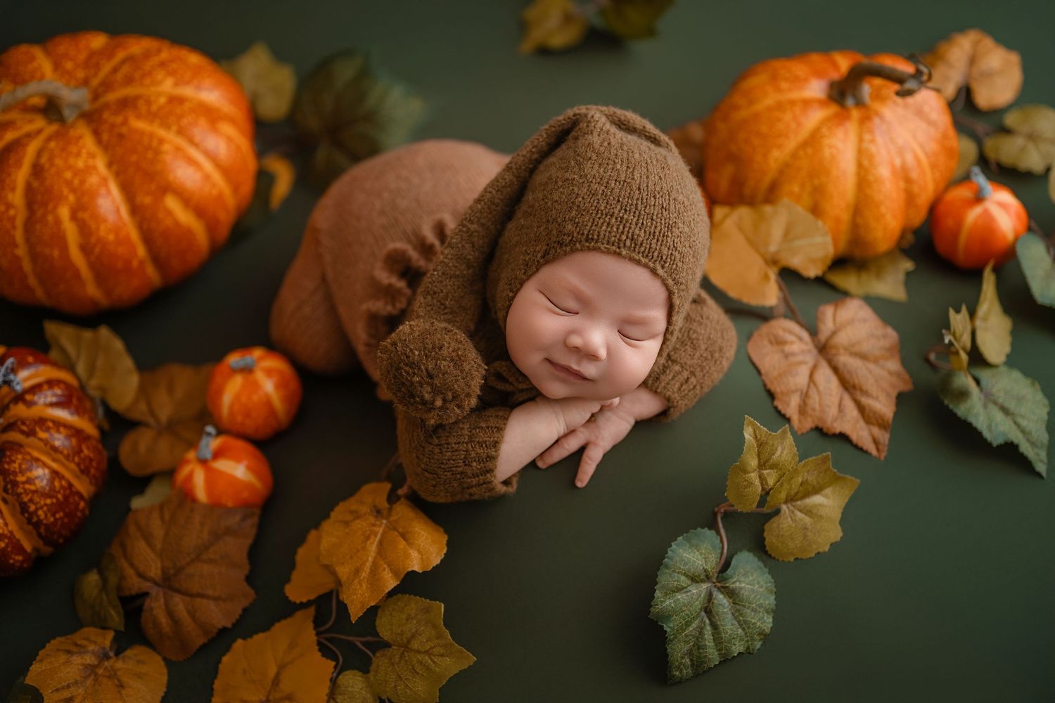 Smiling sleeping newborn in brown knit bonnet nestled among orange pumpkins and autumn leaves.