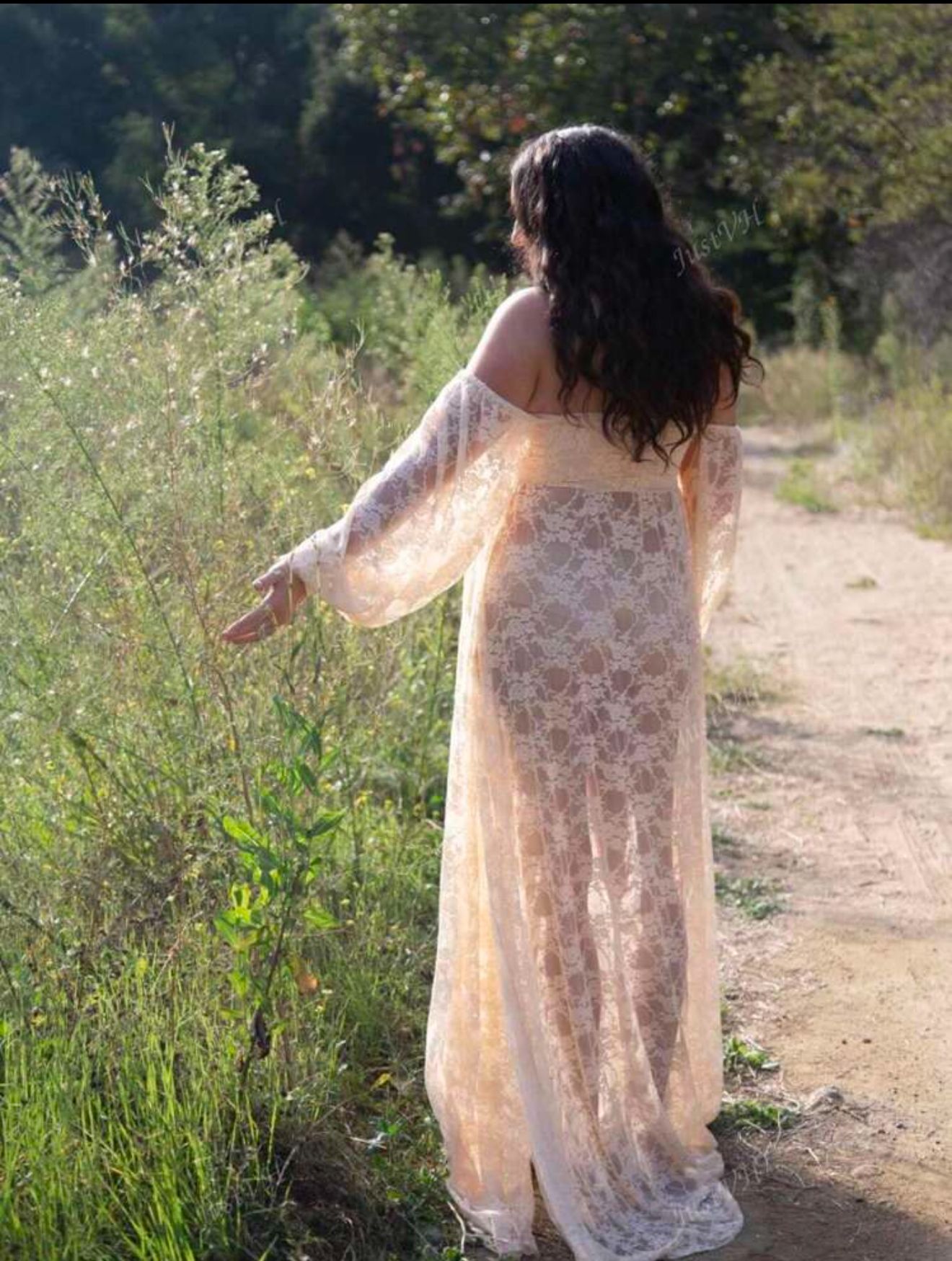Woman in sheer cream lace off-shoulder boho maternity gown walking along a sunlit nature trail.