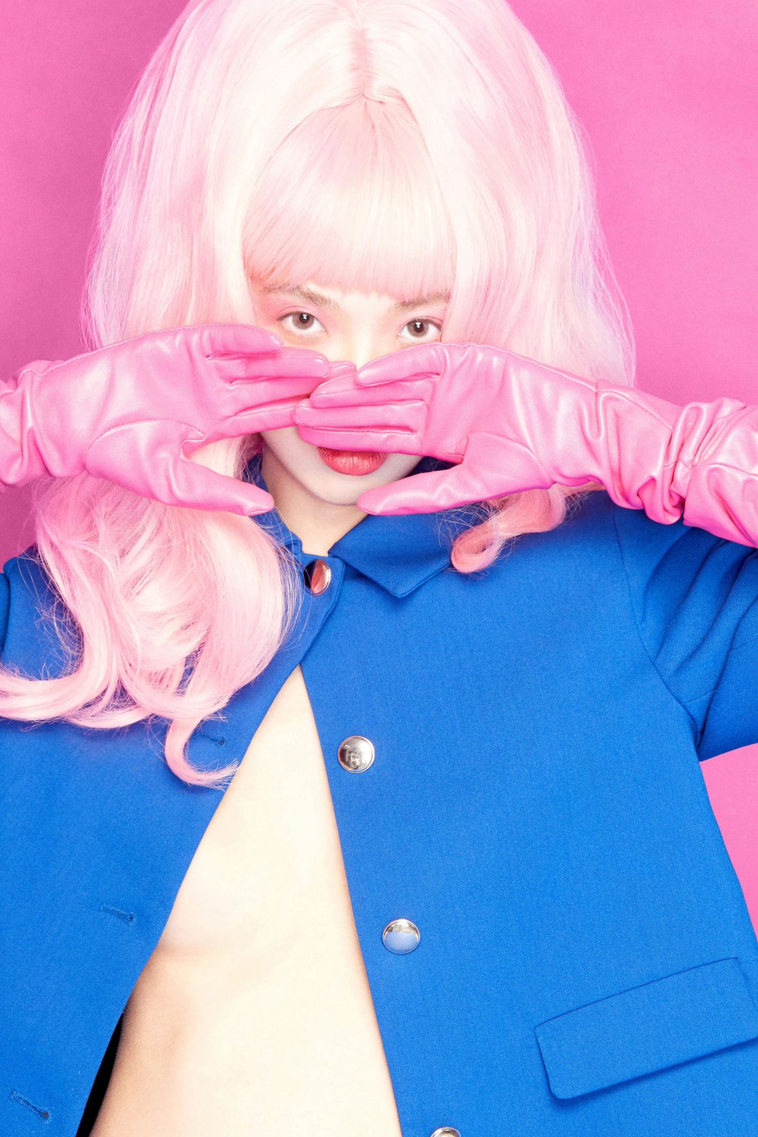 Woman in pink wig and blue jacket poses with pink gloves covering her mouth on a pink background.