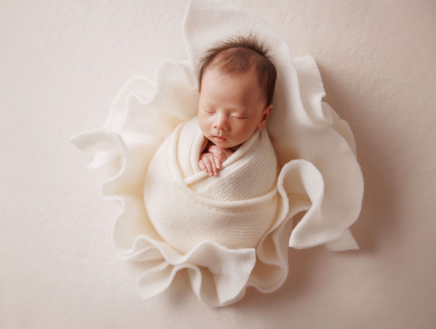 Newborn baby wrapped in cream cloth nestled in a ruffled fabric flower on a soft beige background.