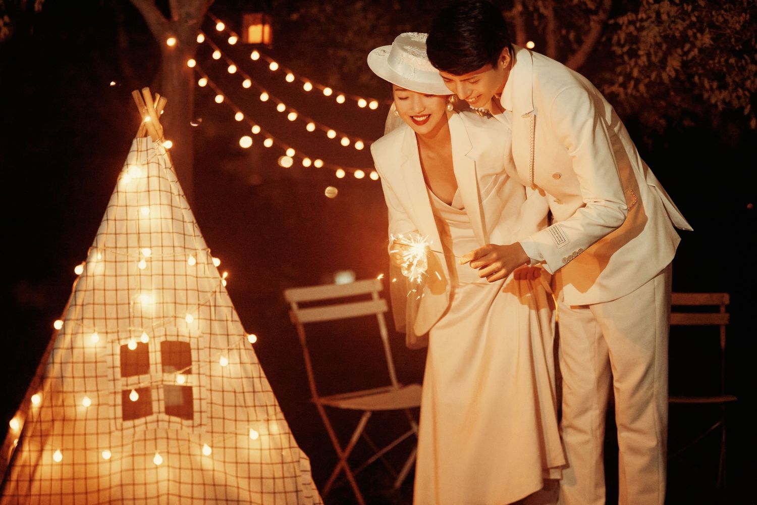Couple in elegant white attire share romantic moment by glowing teepee lantern with string lights at night.