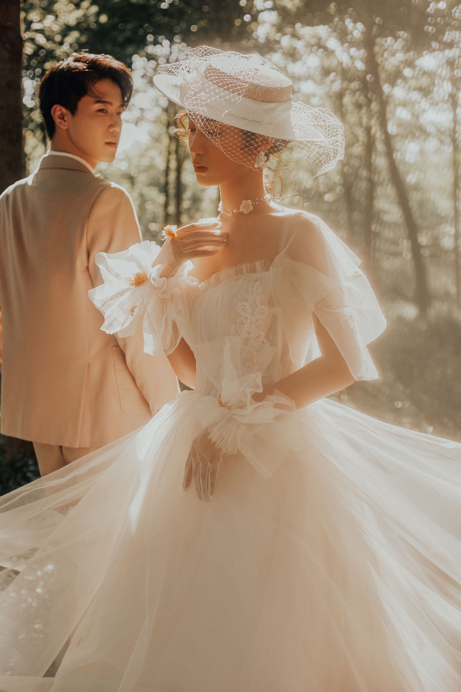 Stunning bride in ornate white tulle ballgown with veiled hat poses with groom in warm sunlit forest setting.