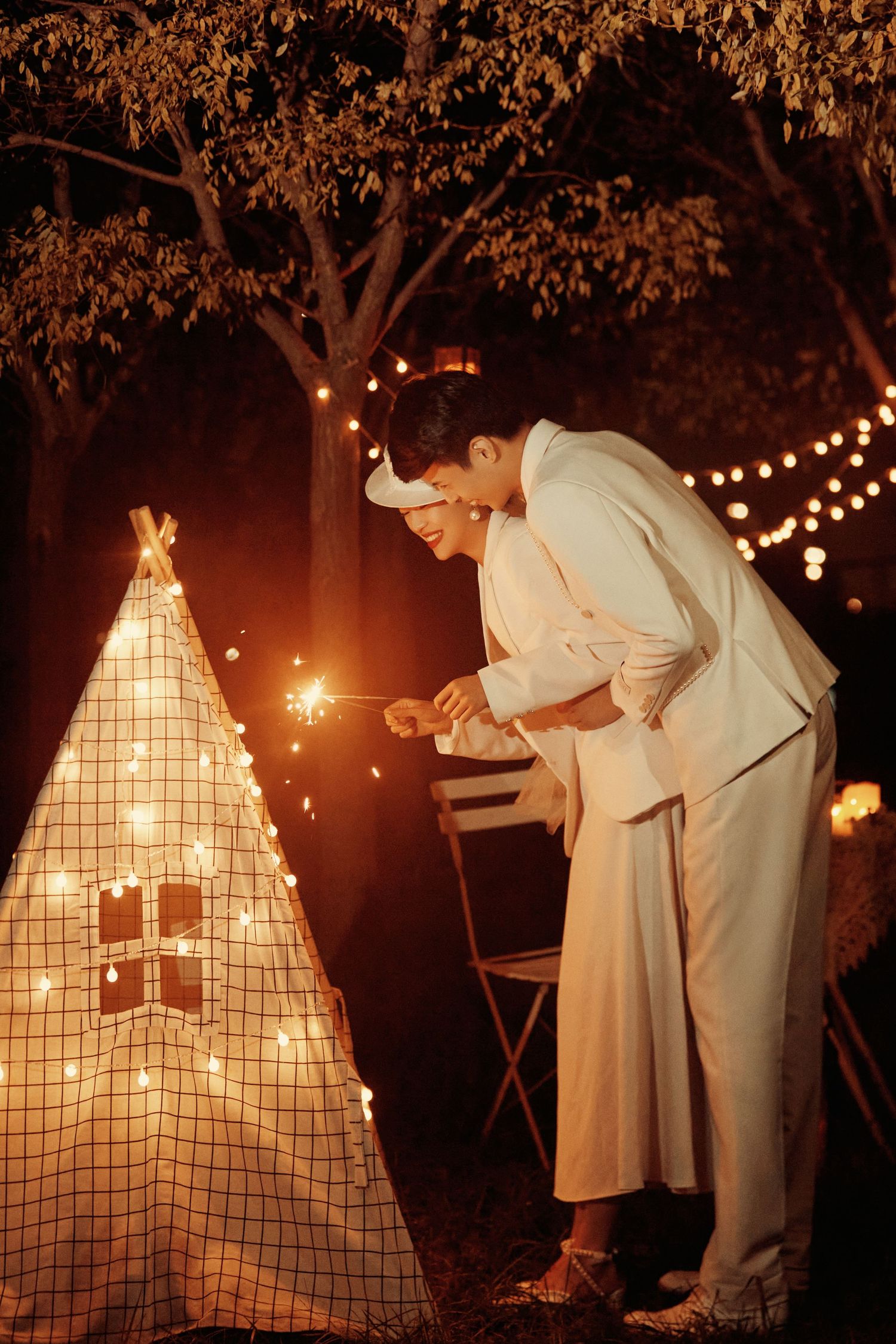 Wedding couple in white attire lighting sparklers near decorated teepee with string lights at night.