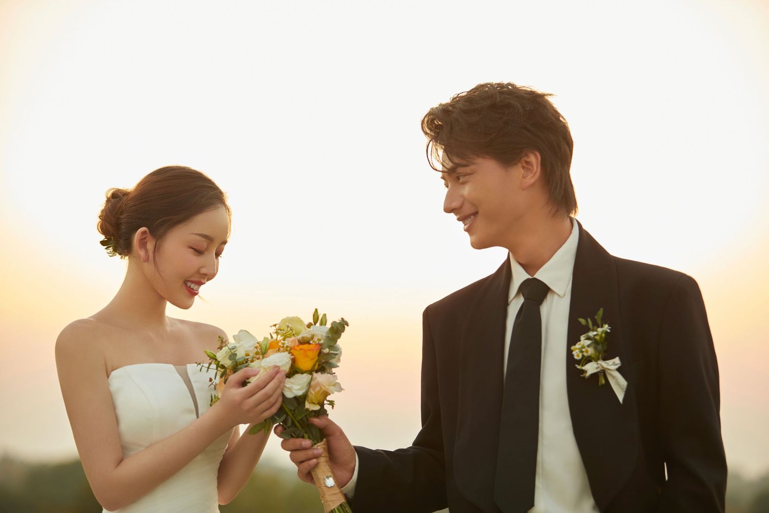 Smiling bride and groom in black suit exchange bouquet at golden sunset outdoor wedding photo.