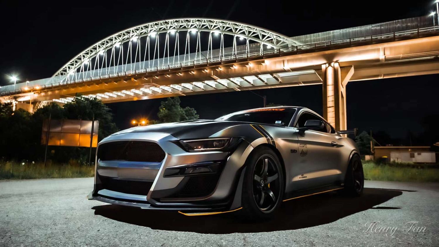 A silver Ford Mustang Shelby GT500 with wide-body kit parked under an illuminated arch bridge at night.