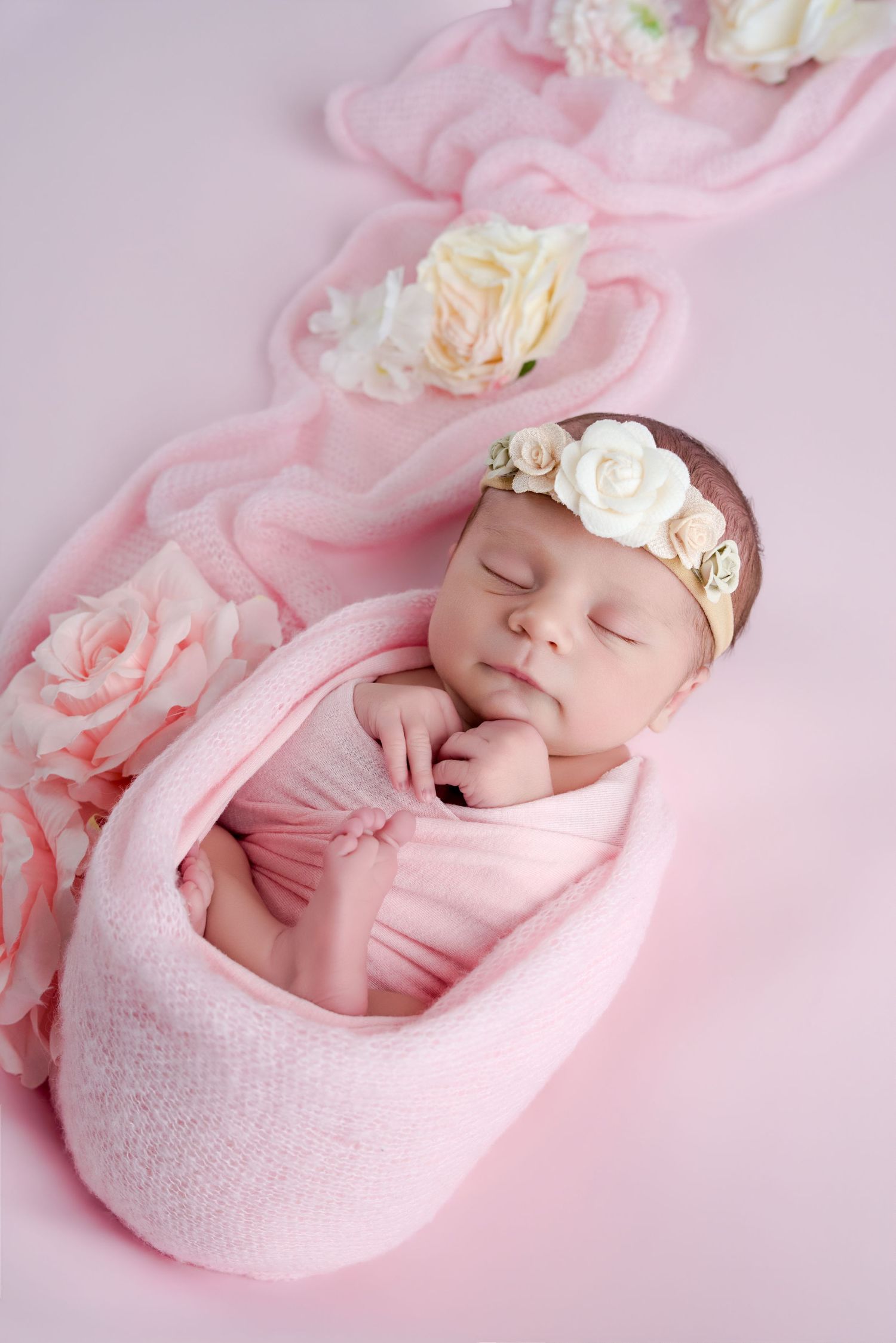 Sleeping newborn baby girl swaddled in pink wrap with floral headband, surrounded by roses on pink background.