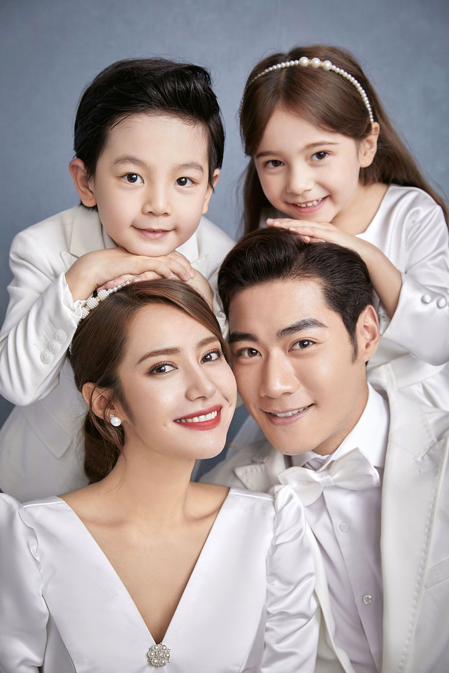 Close-up family portrait of smiling parents in white with two children resting chins on their shoulders.