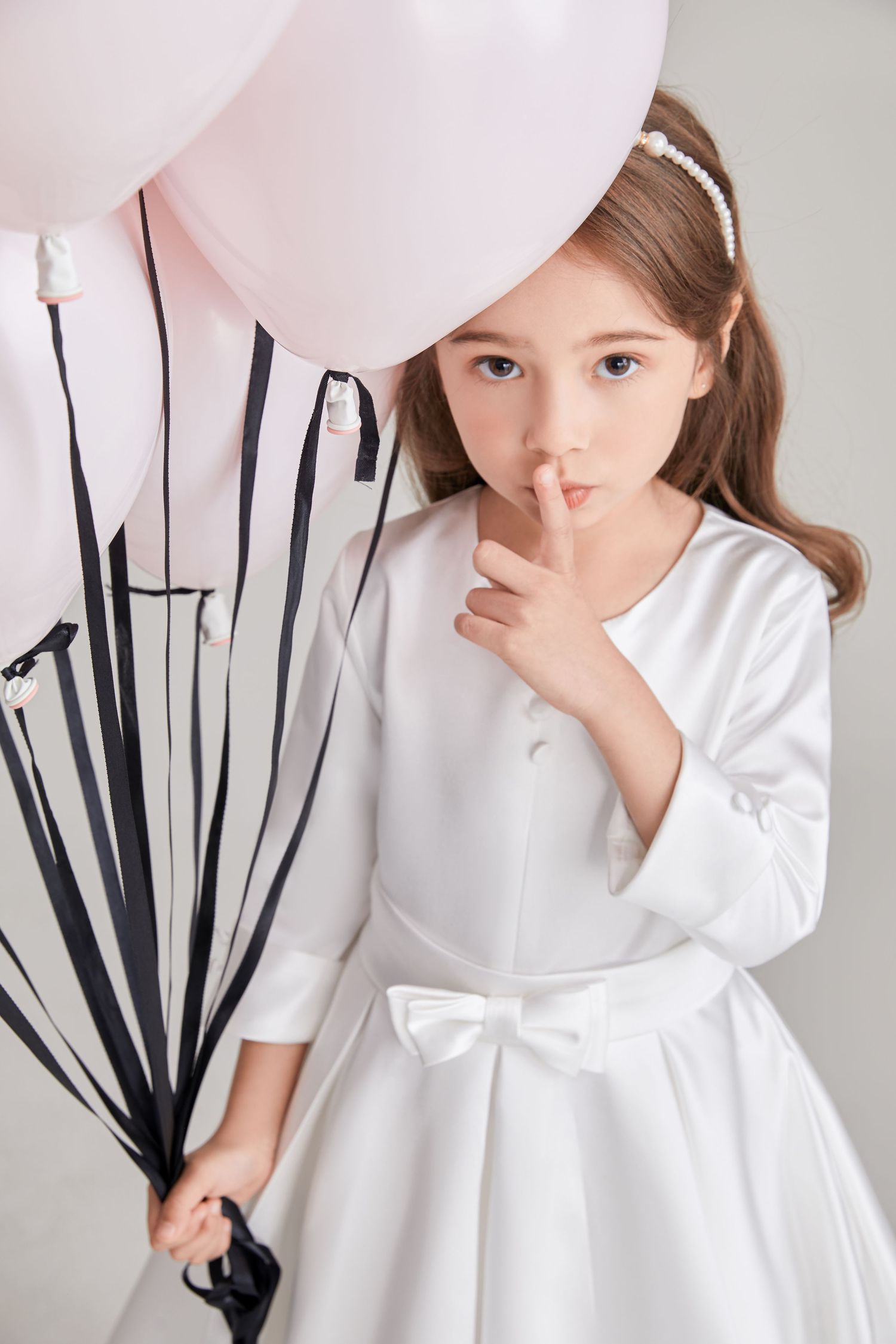 Little girl in white satin dress with bow holds pink balloons and makes shush gesture.