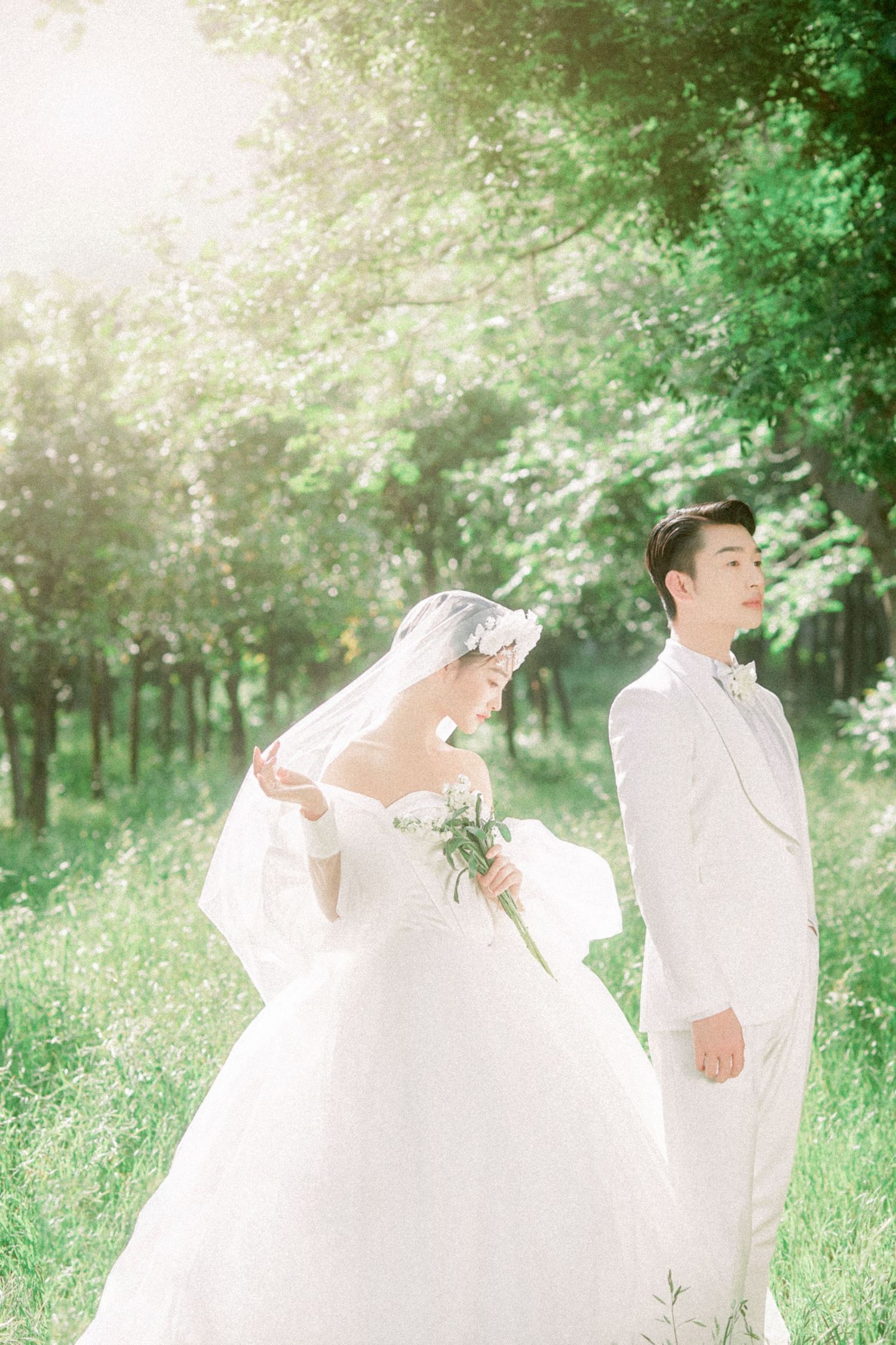 Asian bride and groom in white wedding attire standing in sunlit green forest, romantic outdoor ceremony.