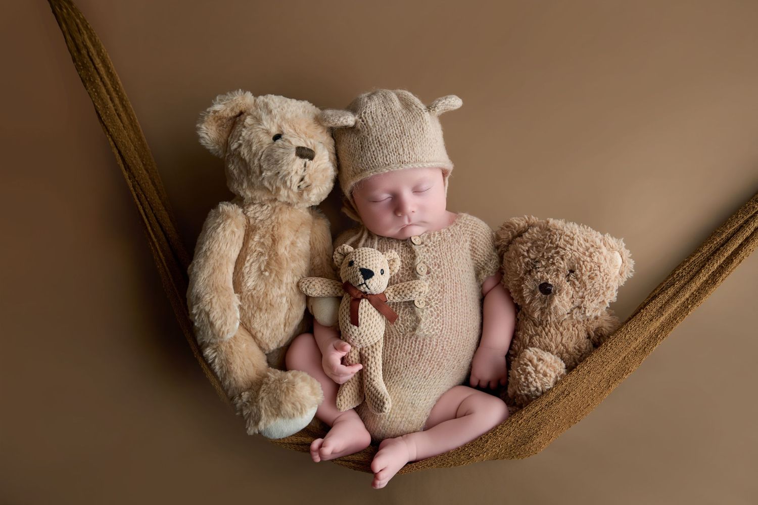 Newborn baby sleeping in a hammock wearing a bear hat, surrounded by three soft teddy bears on a brown background.