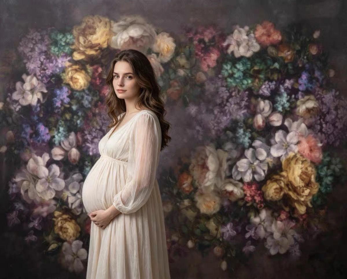 Pregnant woman in white gown poses before floral backdrop with colorful blooms for elegant maternity portrait.