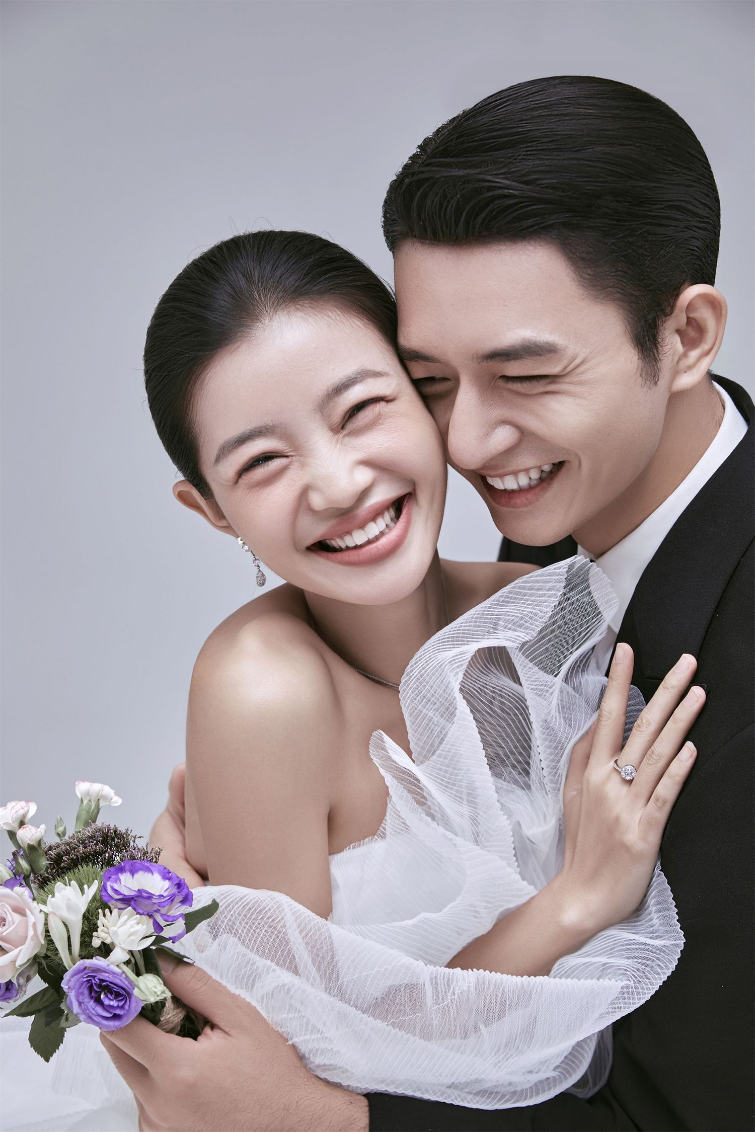 Joyful Asian bride and groom laughing together, bride holding purple floral bouquet in studio.