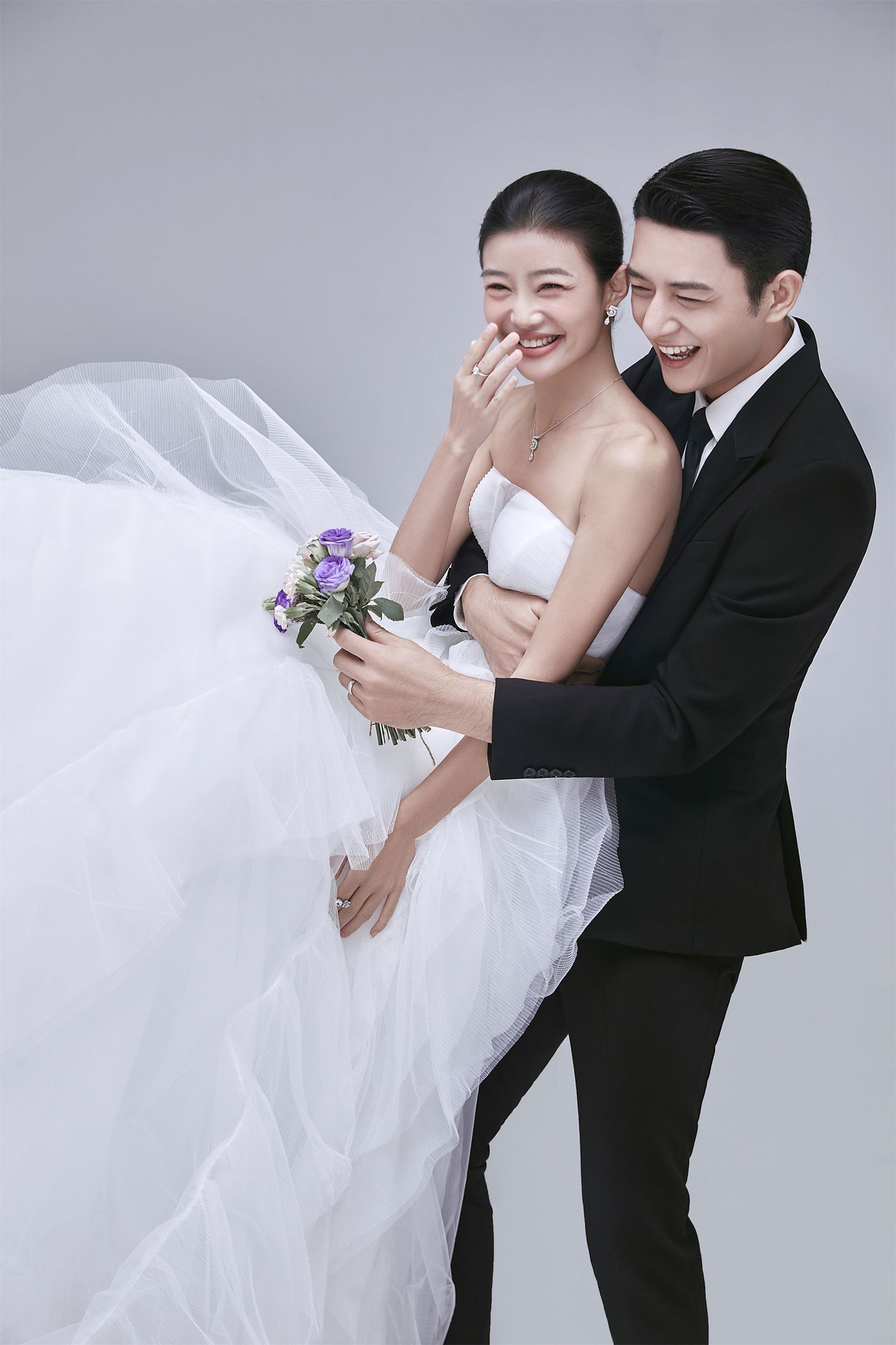 A laughing bride in a strapless white gown is embraced from behind by her groom in a black suit.