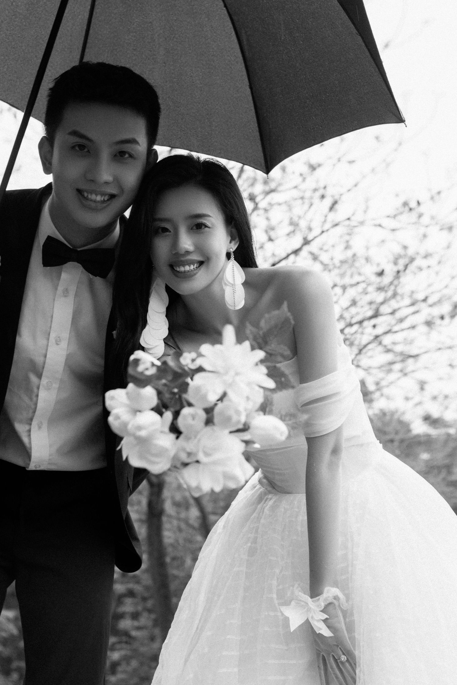 Smiling bride and groom pose together under an umbrella holding a bouquet in a classic black and white wedding photo.