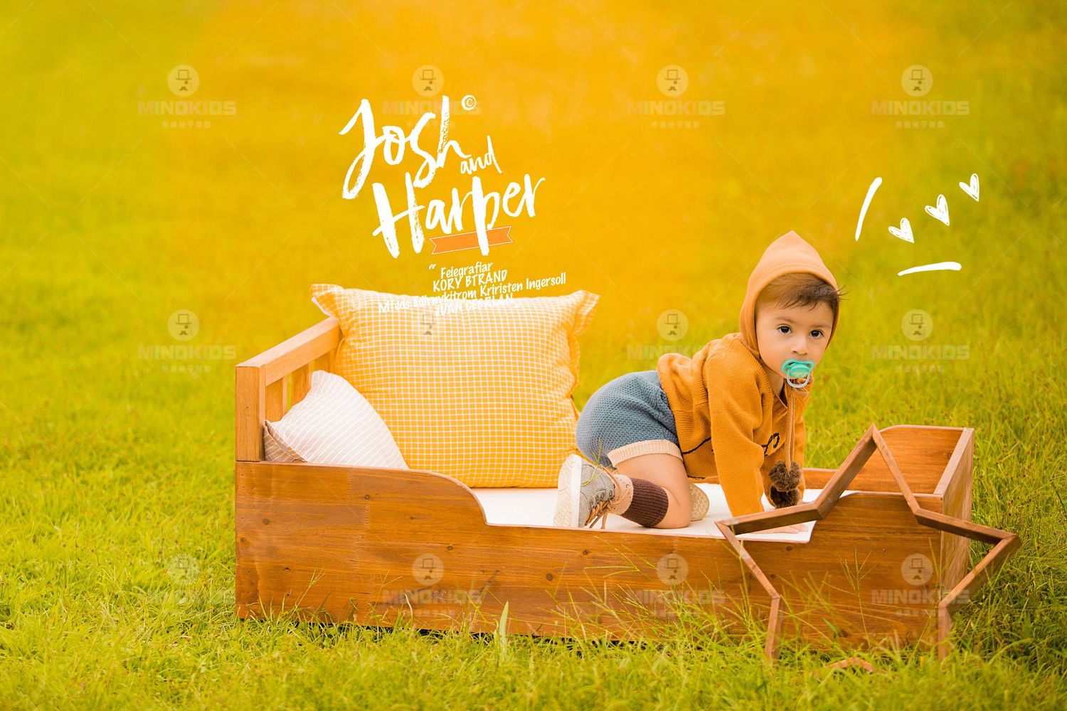 Baby in orange hoodie with pacifier relaxes on wooden bed prop in lush green grass outdoor setting.
