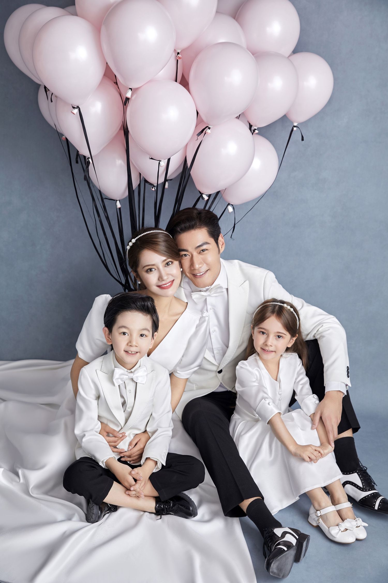 Family of four in white outfits sitting together surrounded by pink balloons in a studio portrait.