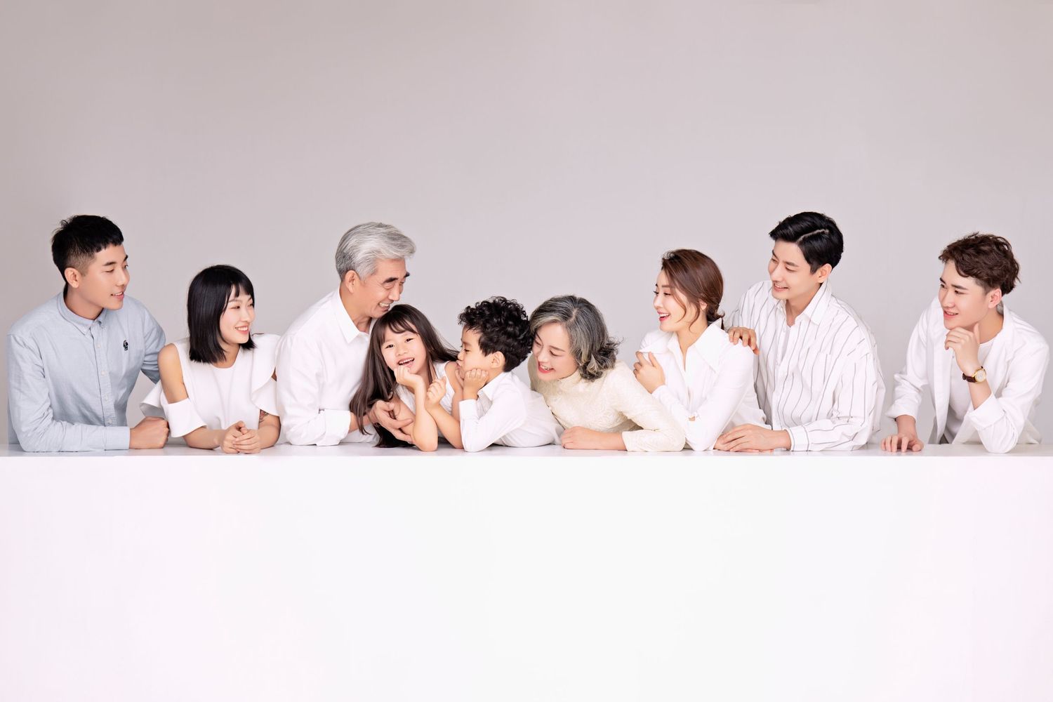 A large multigenerational Asian family in white shirts laughing and leaning on a white surface.
