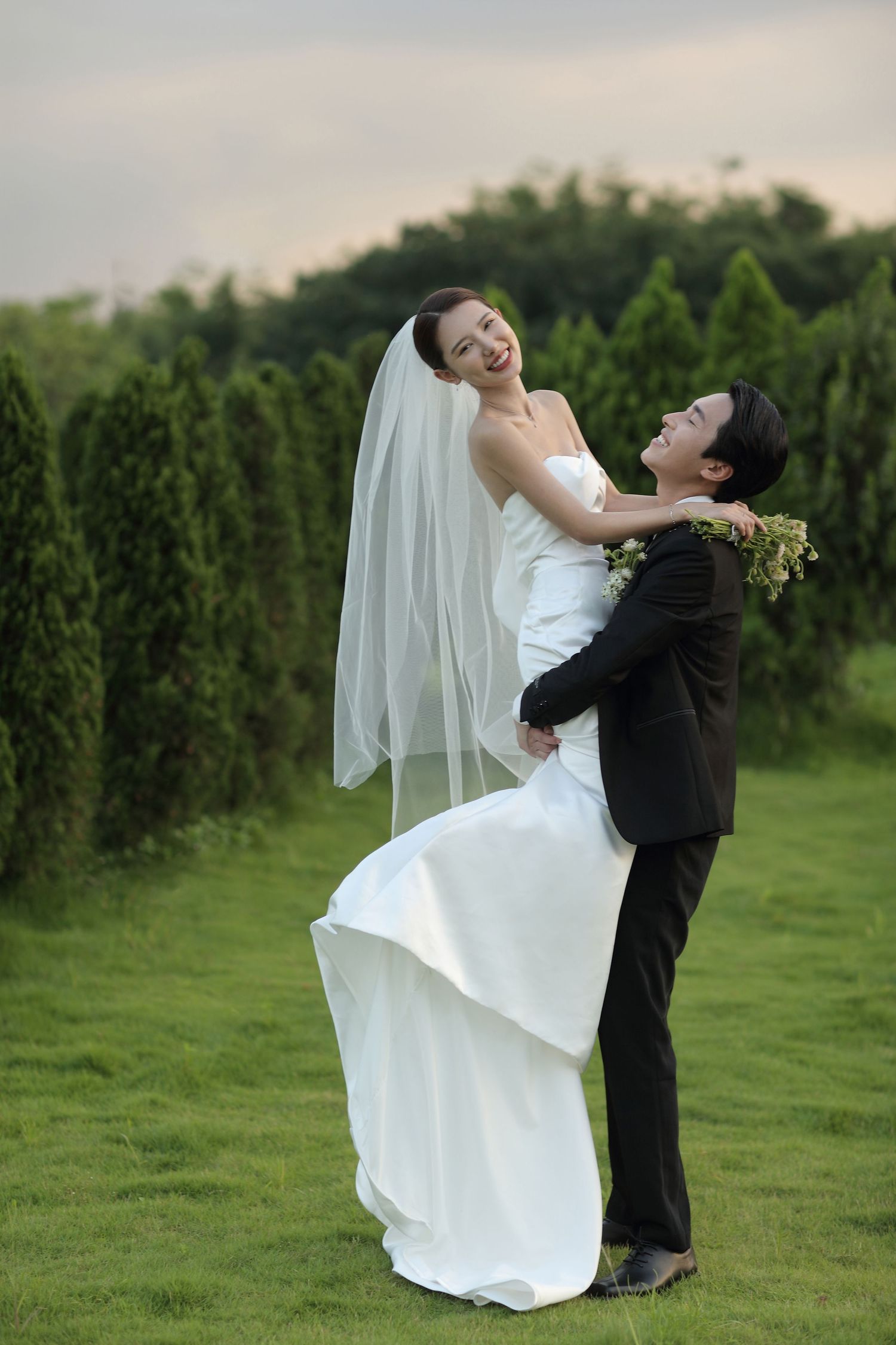 Groom lifting smiling bride in white wedding gown outdoors on green lawn at dusk.