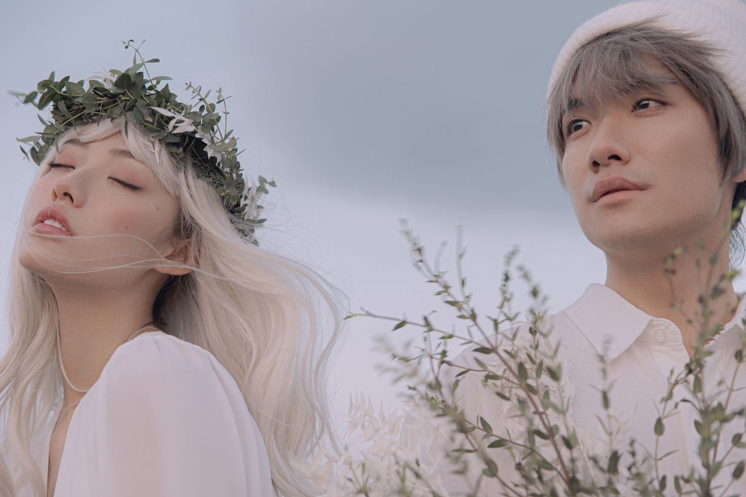 A couple in white outfits with floral crowns pose dreamily among tall white wildflowers under a cloudy sky.