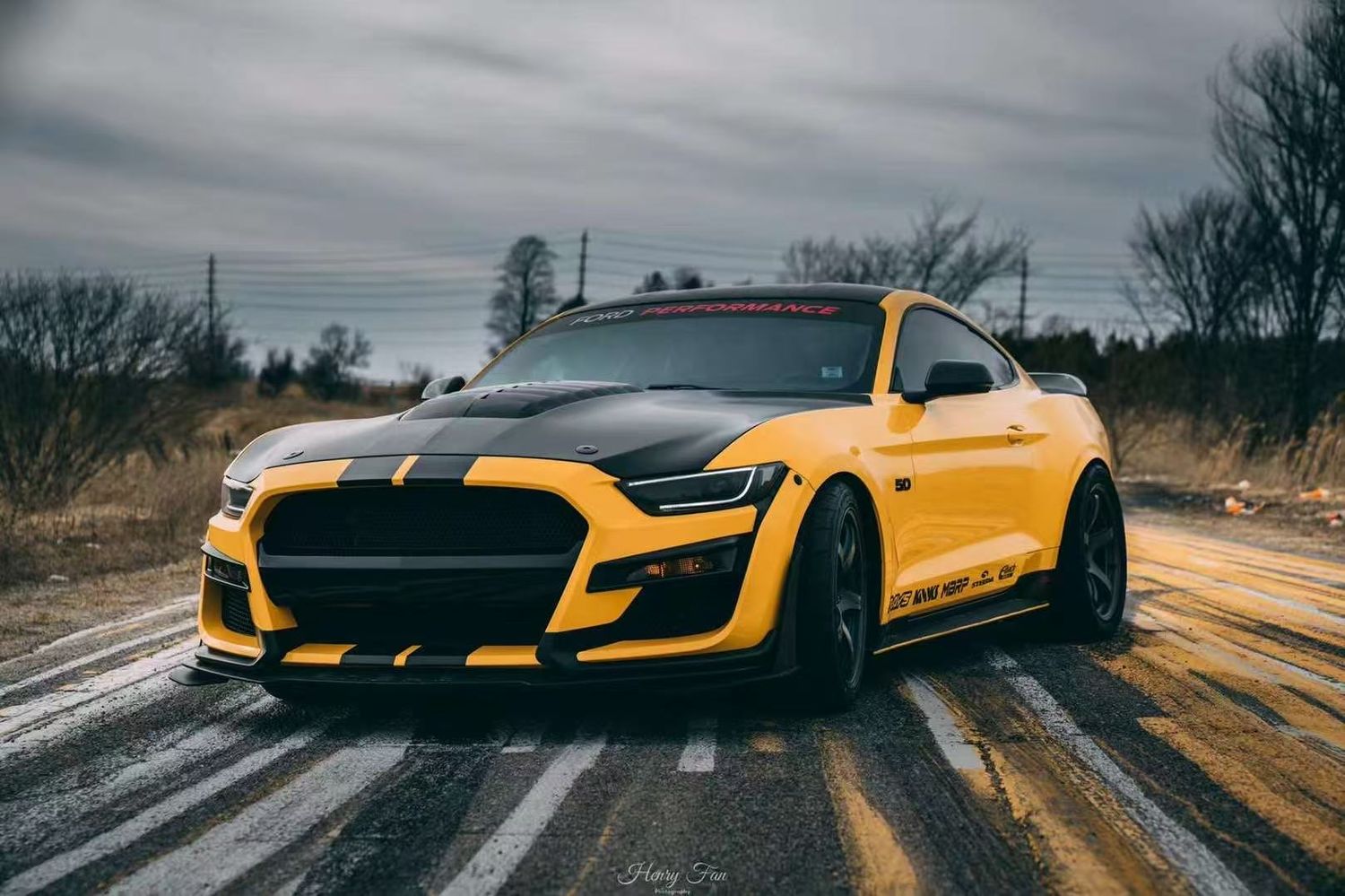 A yellow and black Ford Mustang Shelby GT500 with racing stripes parked on a wet road under cloudy skies.