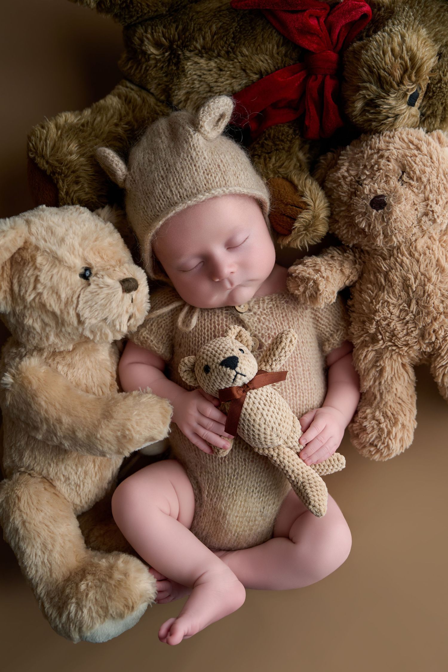 Sleeping newborn baby in knit bear outfit and hat, surrounded by teddy bears, holding a small crocheted bear toy.