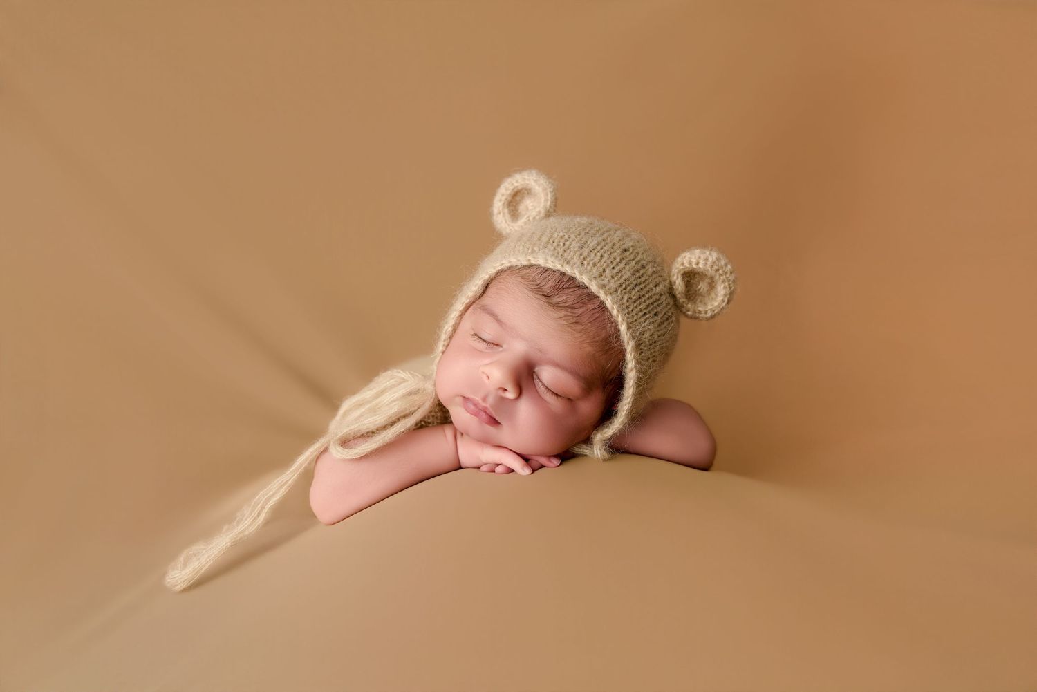 Newborn baby sleeping peacefully wearing a knitted beige bear bonnet hat on a warm tan background.