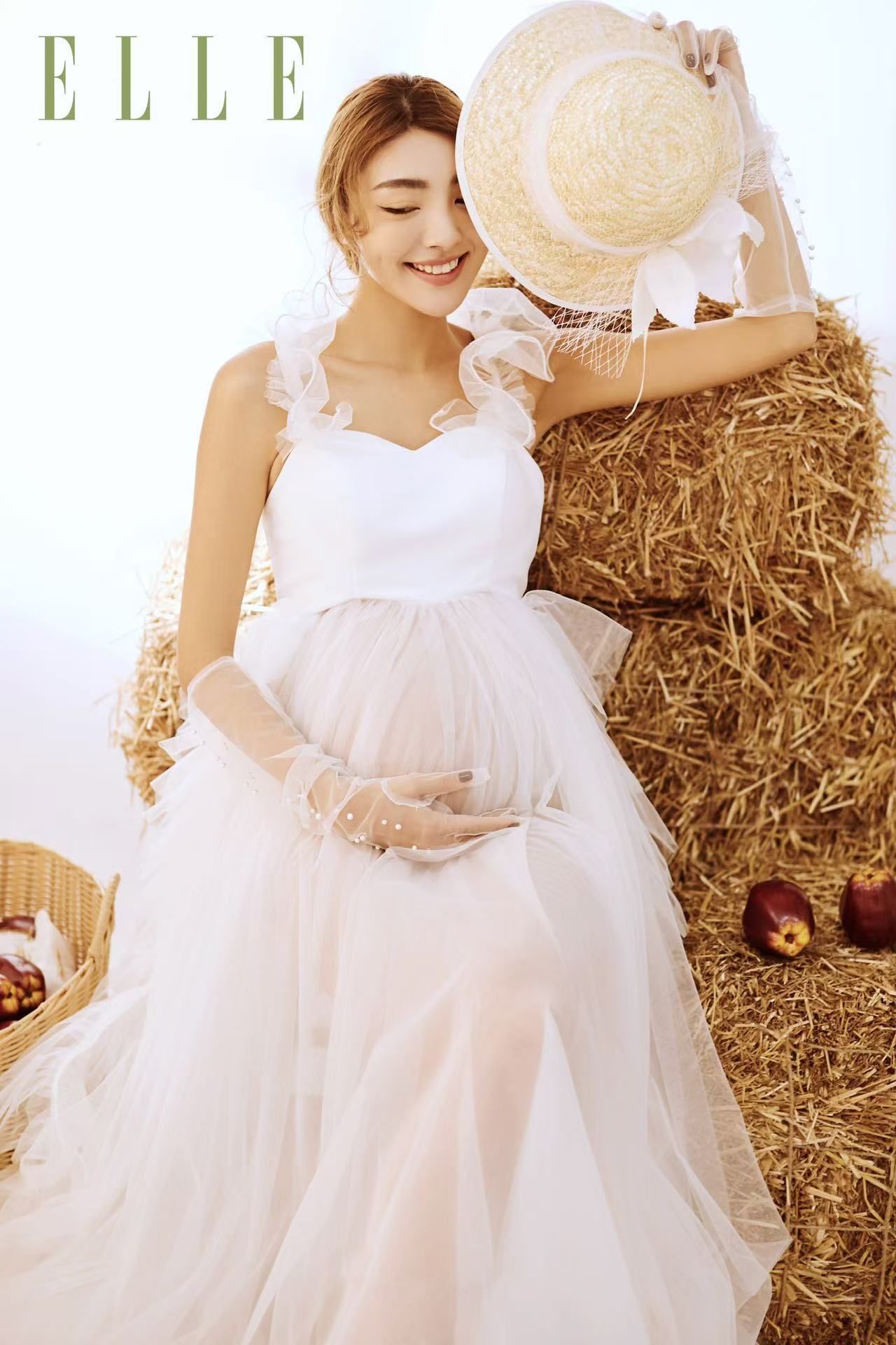 A pregnant woman in a white ruffled tulle gown poses with a straw hat beside hay bales in an ELLE magazine shoot.