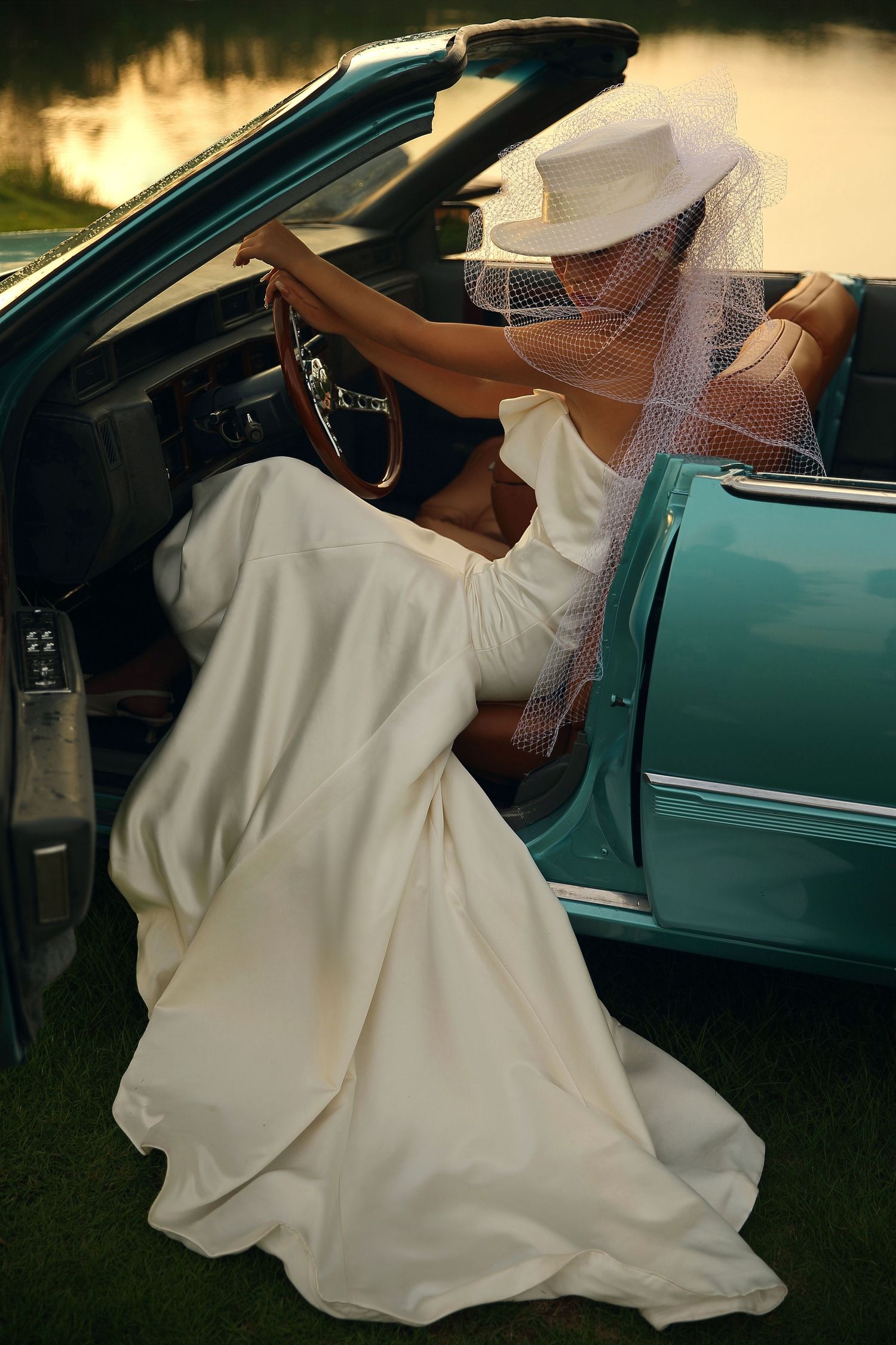 Bride in flowing white gown and veiled hat stepping into a teal vintage convertible at sunset.