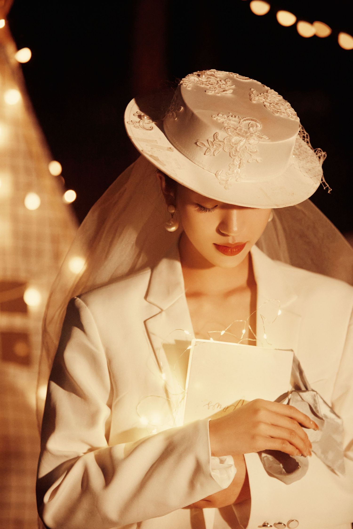 Elegant bride in white blazer and hat reading glowing letter under warm string lights at night.
