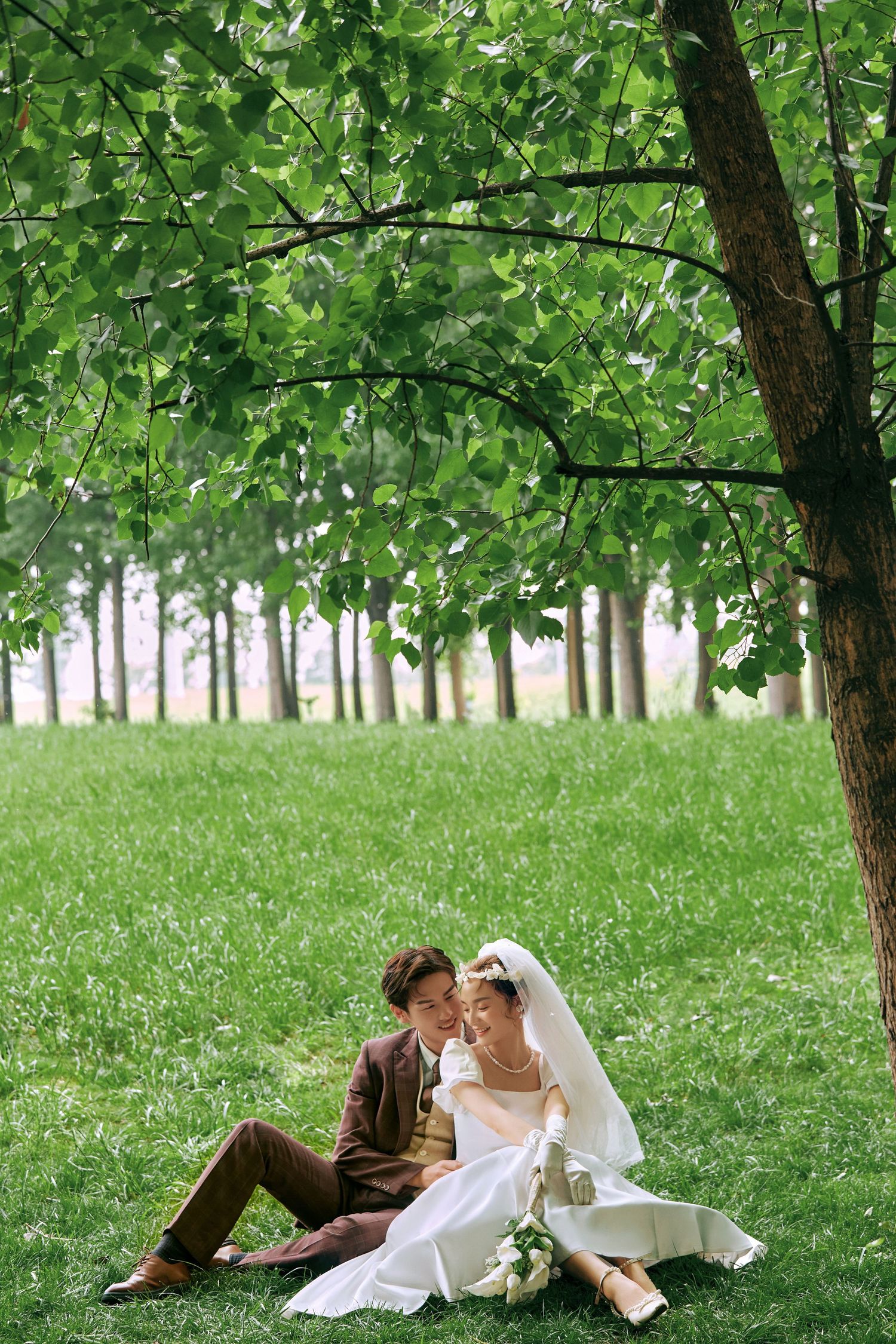 Bride and groom sitting together on grass under large tree in scenic green outdoor setting.