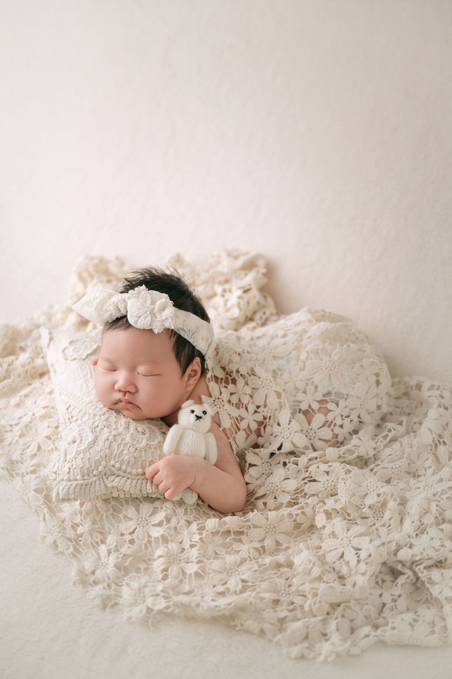 Newborn baby girl sleeping with tiny white teddy bear on lace blanket.