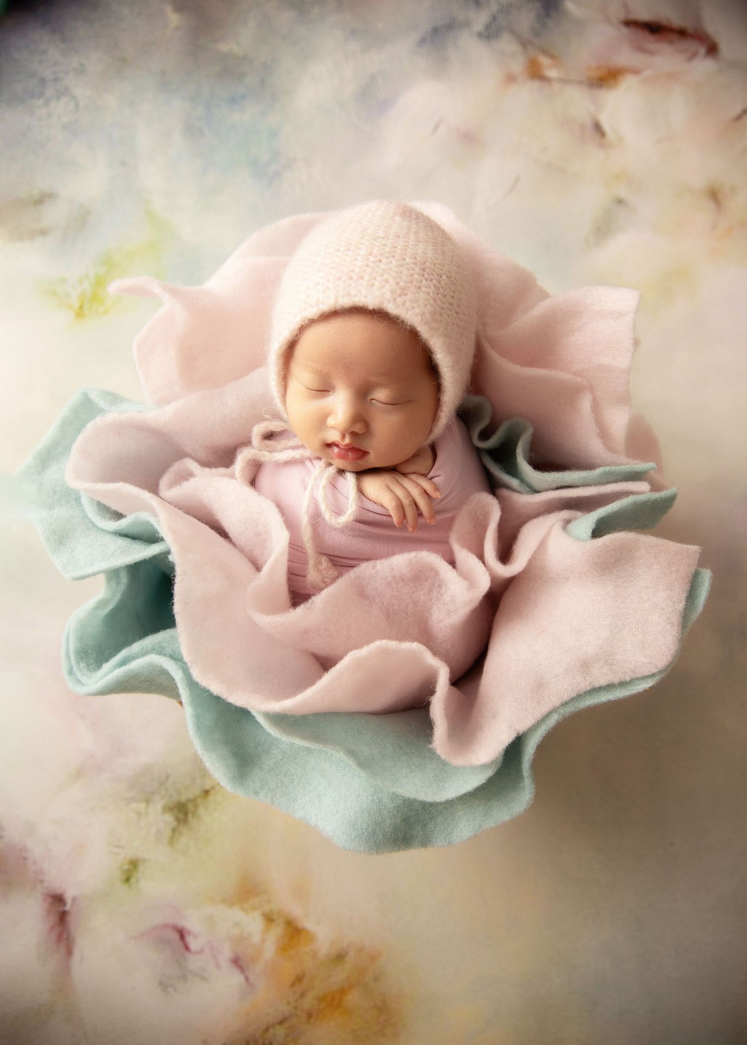Sleeping newborn baby in pink wrap and bonnet nestled in a flower bowl on a soft floral background.