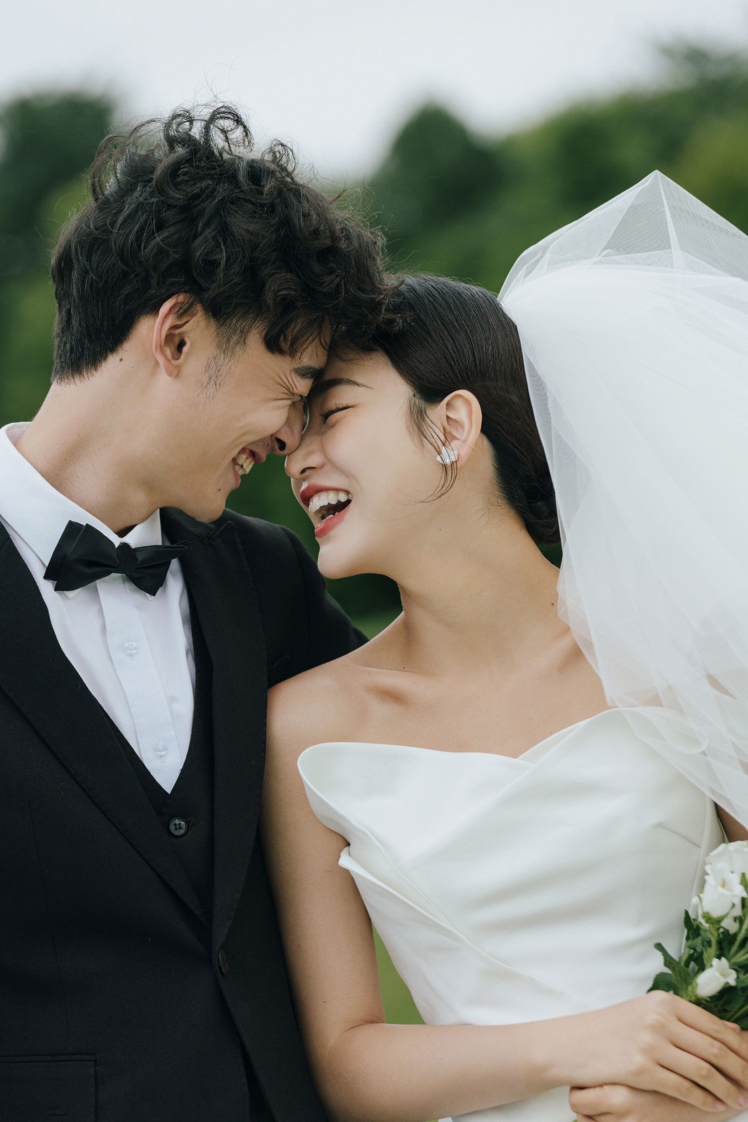 Joyful bride and groom laughing together outdoors on green lawn in elegant wedding attire.
