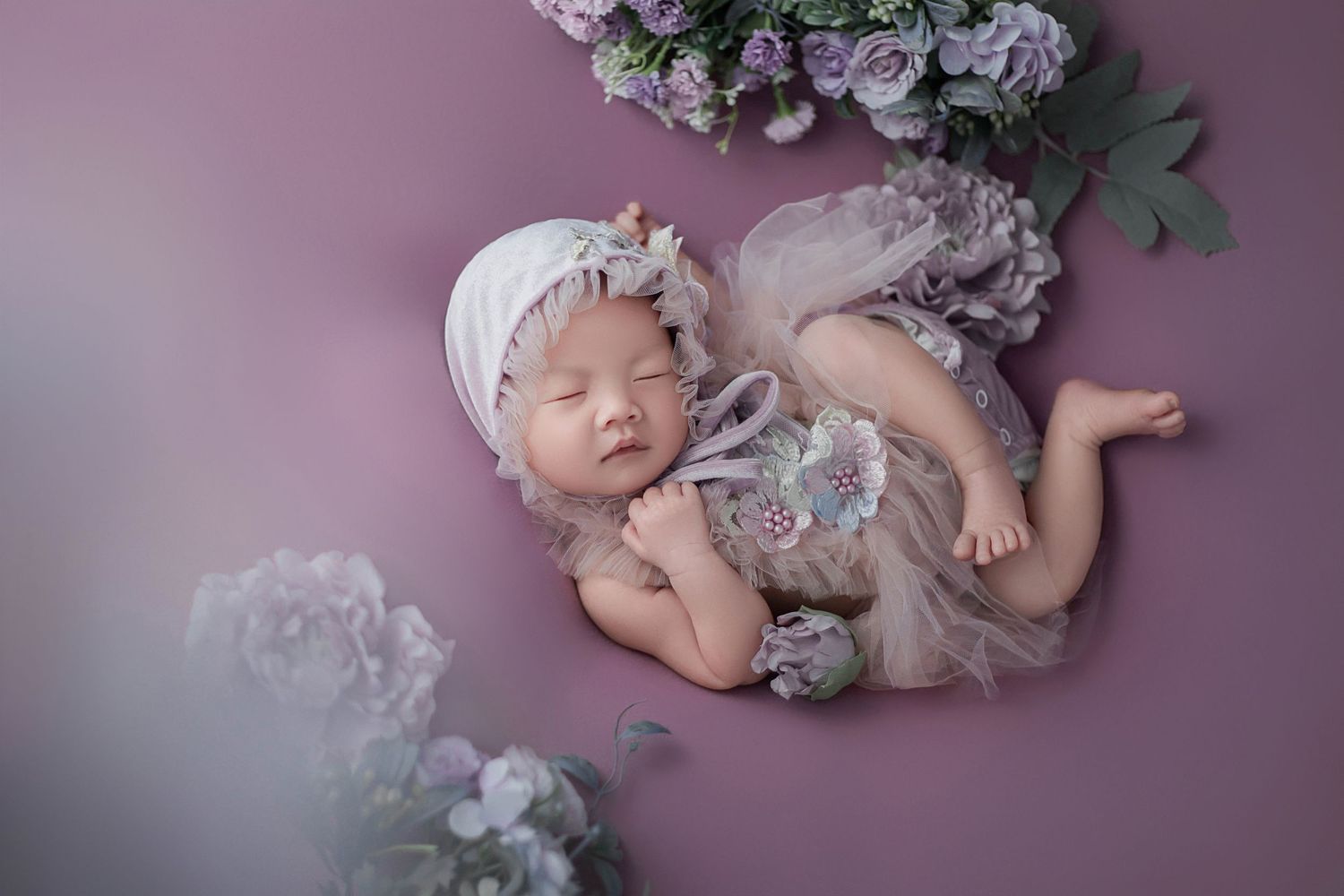 Newborn baby girl sleeping on purple backdrop in lavender tulle outfit surrounded by purple flowers.