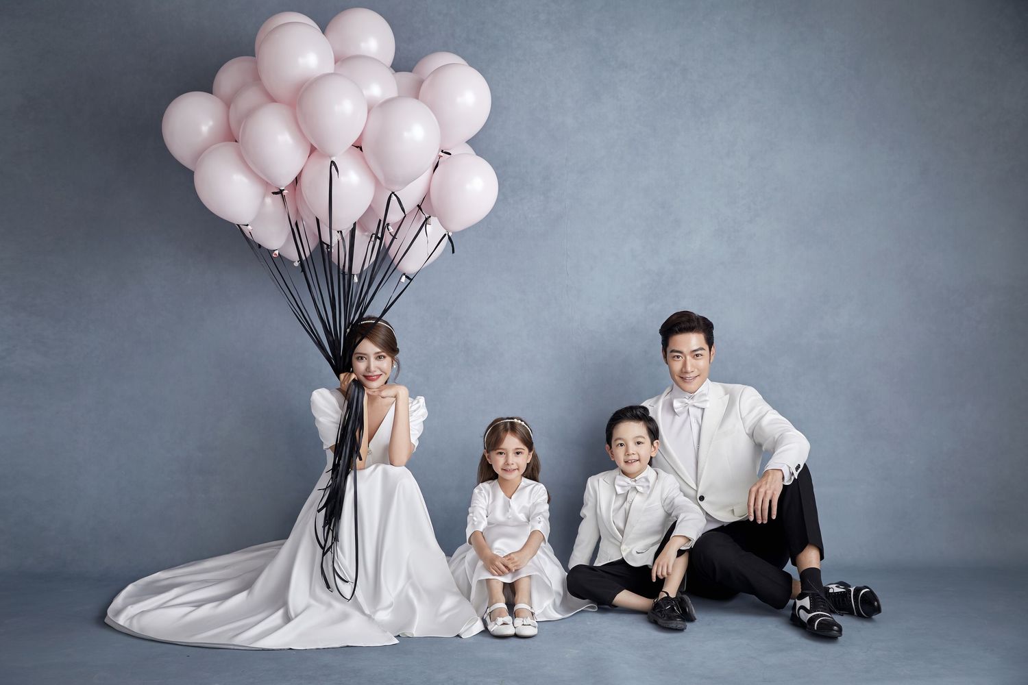 Family of four in white outfits posing with pink balloons in a studio portrait photo.