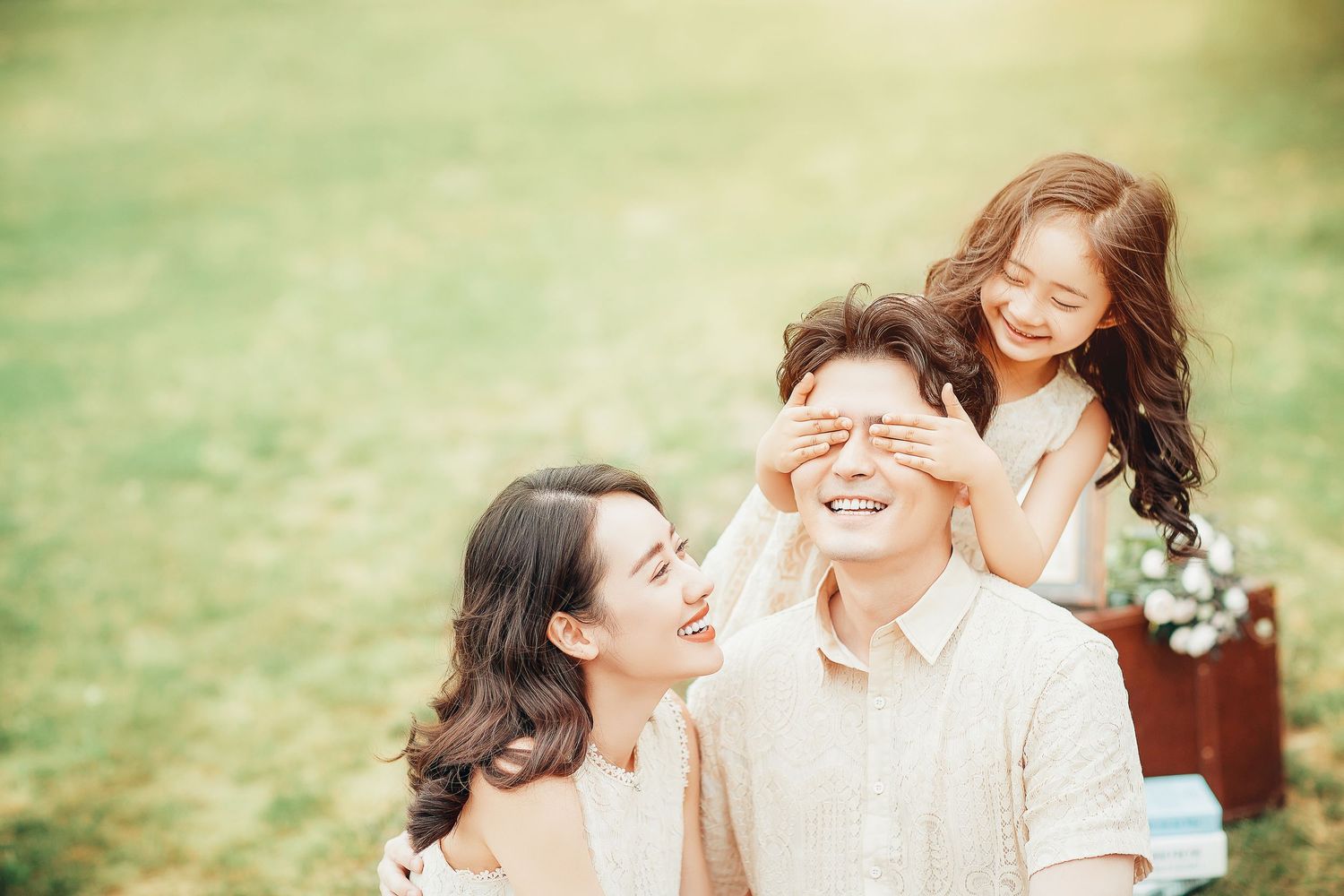 A smiling Asian girl playfully covers her fathers eyes while mother laughs during a park photoshoot.
