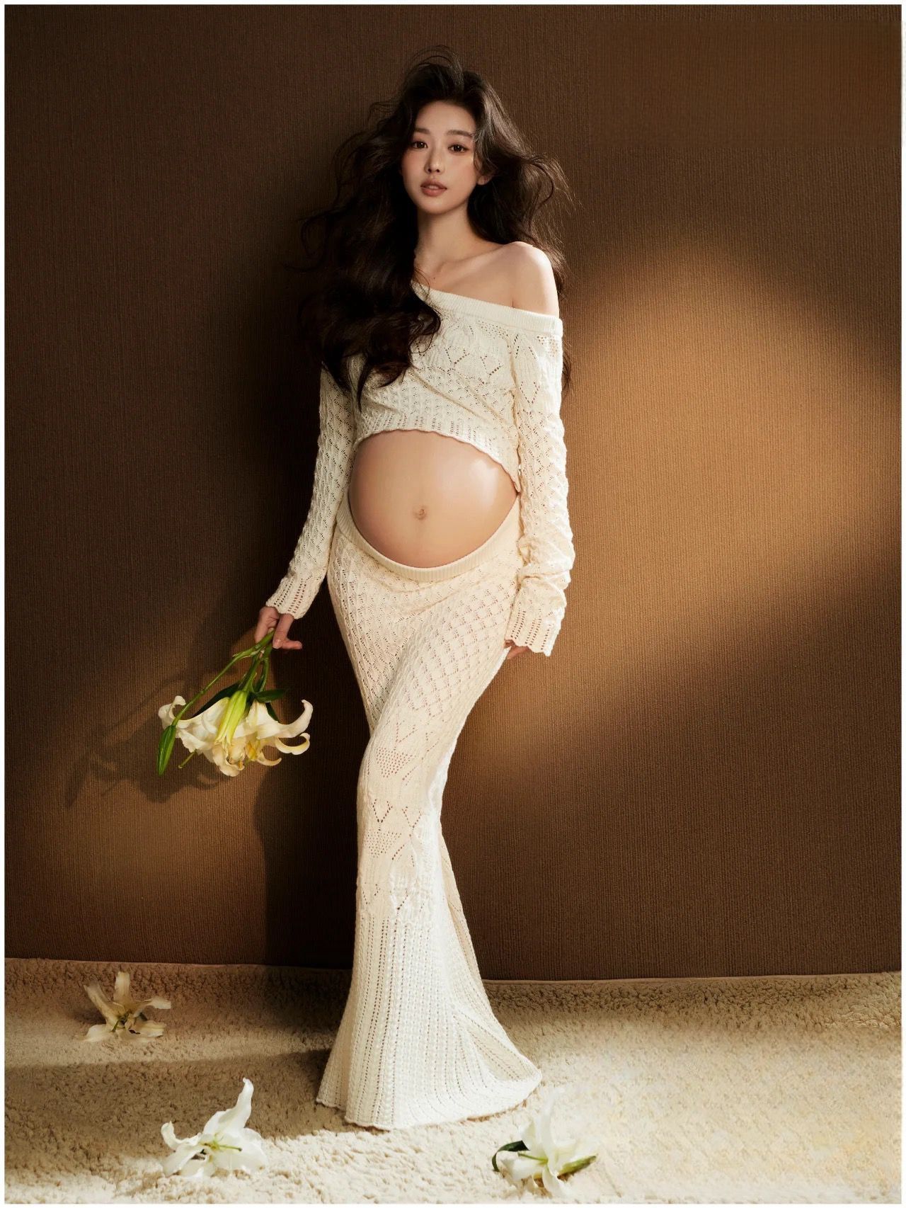 Pregnant woman in cream knit crop top and maxi skirt holding white lilies in elegant maternity photo shoot.