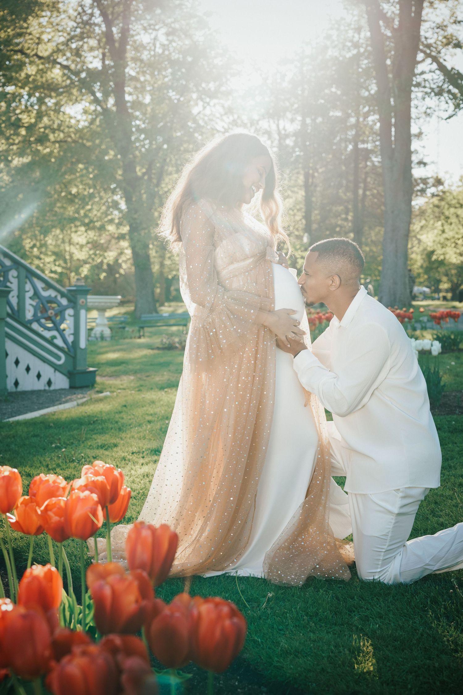 Man in white suit kneeling to kiss pregnant woman's belly in golden gown among red tulips in sunlit garden.